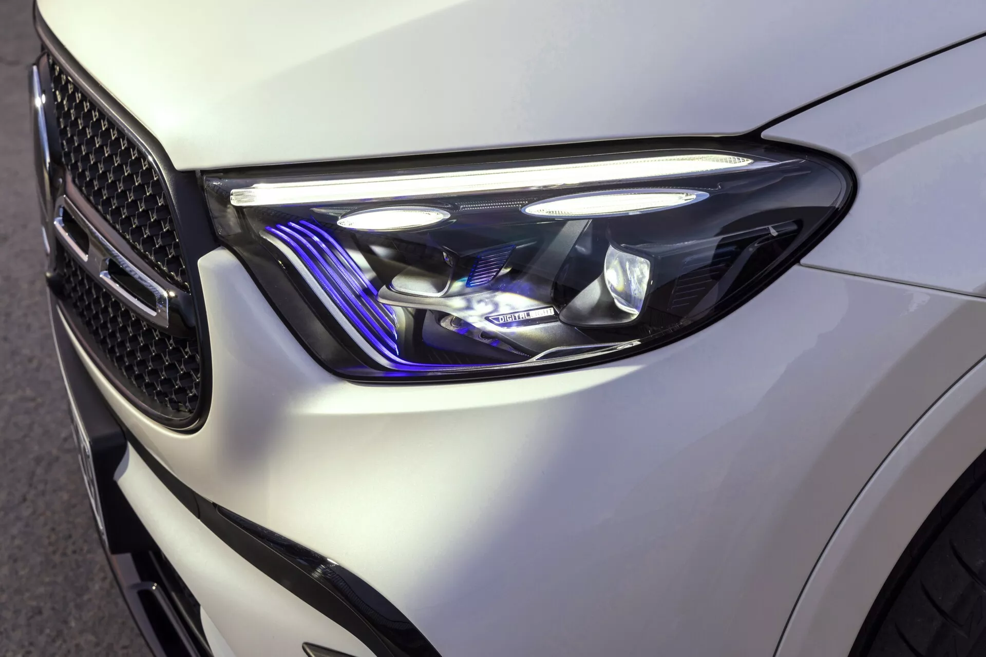 Gros plan sur le quart avant gauche d'une Mercedes GLC 2022, mettant en valeur le design moderne et élégant des phares avec des formes angulaires nettes et des éléments d'éclairage sophistiqués, notamment des feux de jour à LED et un accent bleu. La peinture blanche brillante accentue les lignes bien définies du véhicule et la calandre visible en nid d'abeille reflète son design orienté vers la performance.
