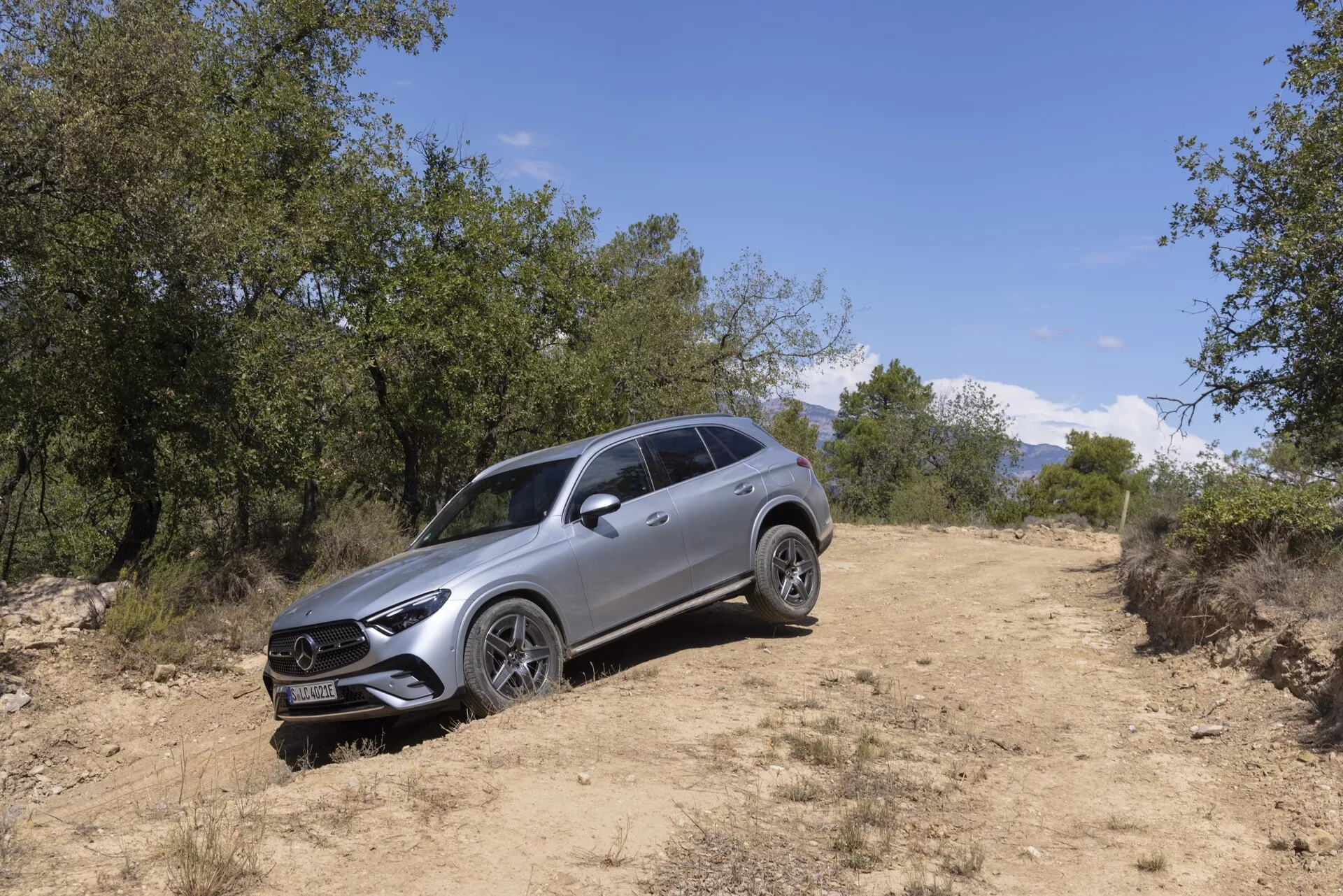 Image d'un SUV Mercedes GLC 2022 dans un cadre tout-terrain prise selon un angle trois quarts de l'avant gauche. Les caractéristiques visibles incluent une grille proéminente avec emblème centré, des phares à LED avec feux diurnes, des jantes en alliage à plusieurs branches, des barres de toit, une carrosserie sculptée, des rétroviseurs avec clignotants intégrés et une garde au sol élevée indicative des capacités tout-terrain. Le véhicule est sur un chemin de terre entouré d'herbe et d'arbres, avec la lumière naturelle du soleil illuminant son design moderne et ses caractéristiques de luxe.