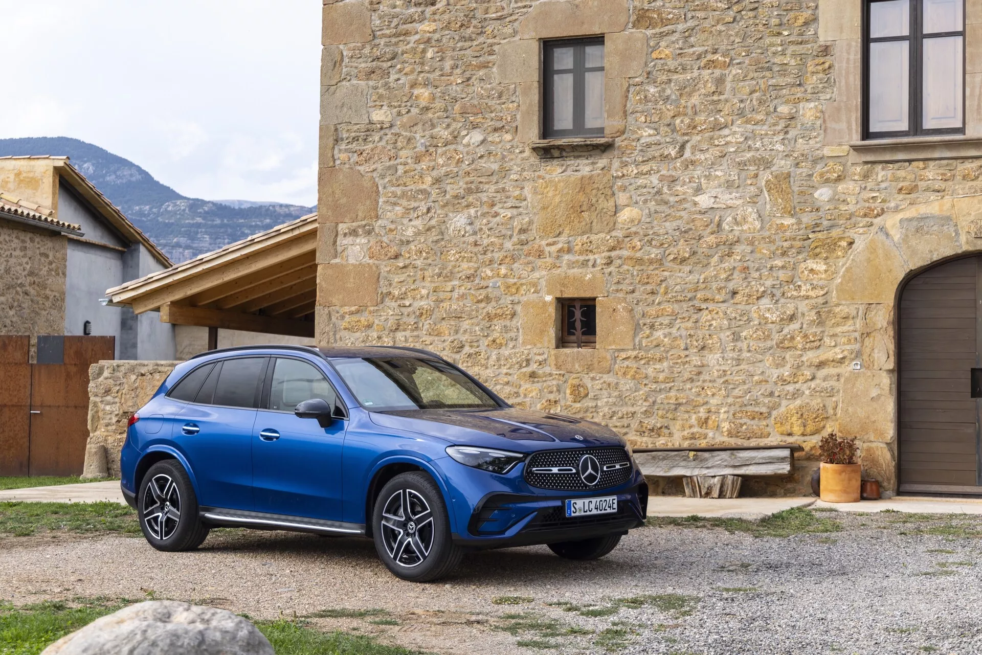 Un SUV Mercedes GLC 2022 bleu moderne est affiché de manière proéminente en plein air, positionné à un angle légèrement décalé mettant en valeur le quart avant droit et une partie du profil latéral. La voiture est ornée d'une calandre avant proéminente avec l'emblème du constructeur, des phares LED horizontaux élégants et de grandes roues en alliage multi-rayons. Elle est située devant un ancien bâtiment en pierre, offrant un contraste saisissant. Les caractéristiques technologiques potentielles telles que le régulateur de vitesse adaptatif, l'assistant de maintien de voie et le toit ouvrant panoramique sont suggérées par son design. La composition de l'image offre une perspective claire et naturelle des attributs luxueux et high-tech du véhicule, capturant l'extérieur détaillé dans un cadre rustique.
