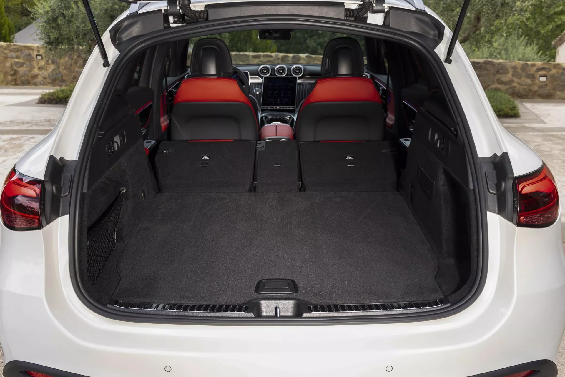L'image montre l'intérieur arrière du Mercedes GLC 2022 (254) avec le hayon ouvert. Le point de vue offre une vue claire de l'espace de chargement spacieux et en moquette, avec un plancher de chargement plat qui met en lumière la flexibilité de l'espace grâce aux sièges arrière rabattus en configuration 60/40. Les intérieurs présentent des sièges en cuir rouge premium, des appuie-tête avec inserts rouges et une continuité de design. Partiellement visibles, le tableau de bord et la console centrale suggèrent des fonctionnalités supplémentaires telles que systèmes d'infodivertissement et contrôle climatique. Le hayon laisse penser à un design convivial pouvant inclure des technologies telles que l'ouverture mains libres ou motorisée. Aucune couverture de coffre n'est présente, indiquant qu'elle pourrait être rétractée ou enlevée.