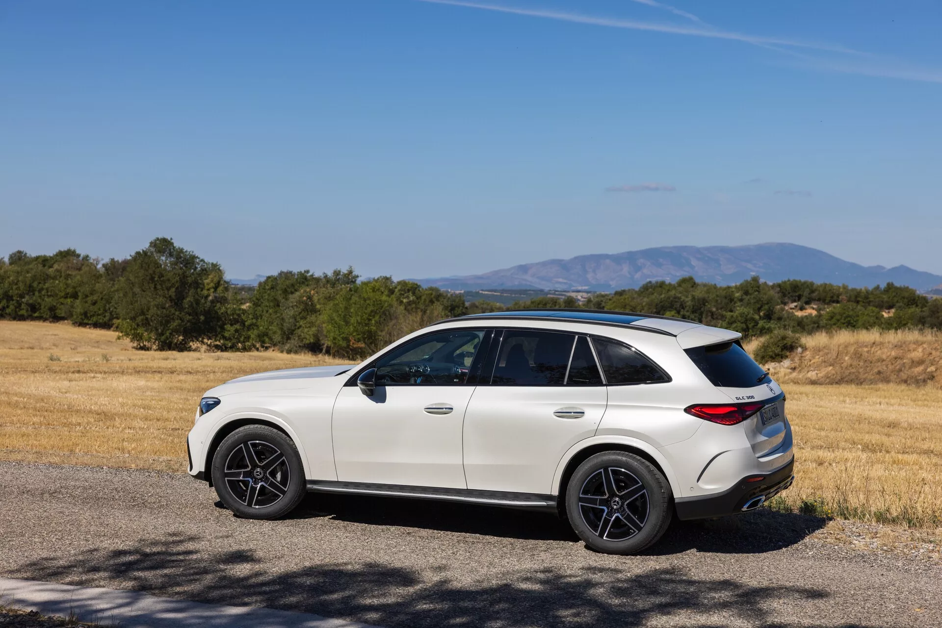 SUV crossover moderne de luxe en milieu rural : vue arrière latérale trois-quarts d'un Mercedes GLC 2022 (254) de couleur claire avec des roues foncées au design sportif, des vitres teintées, une ligne de toit inclinée et un pare-chocs arrière sculpté, garé près d'un champ sec et des arbres par une journée ensoleillée.