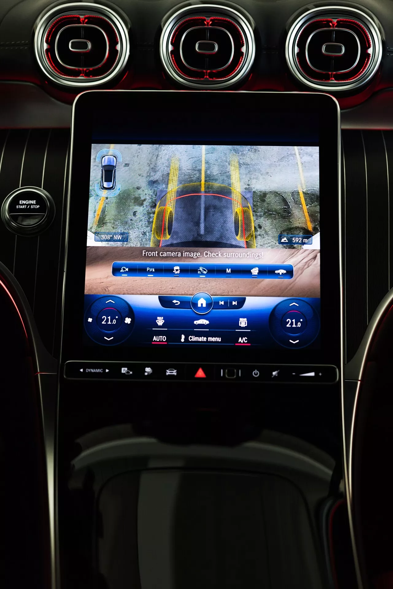 Intérieur du Mercedes GLC 2022 avec console centrale et écran tactile d'infodivertissement affichant le flux de la caméra avant avec des lignes directrices jaunes et bleues pour l'assistance au stationnement. Vue depuis le siège du conducteur comprenant des bouches d'aération avec des détails rouges au-dessus de l'écran et des boutons de contrôle de température en dessous.
