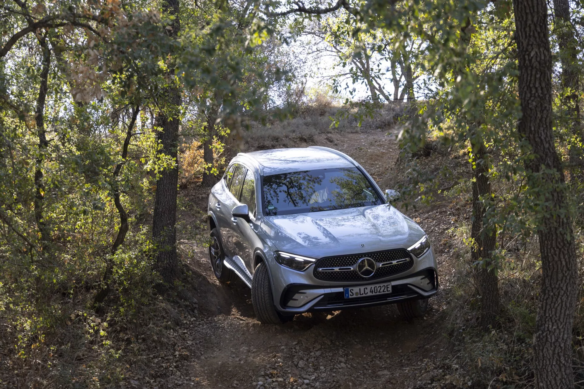 Vue complète du SUV Mercedes GLC 254 de 2022 depuis une perspective frontale trois-quarts, approchant de la caméra sur un sentier naturel entouré d'arbres. L'angle légèrement élevé met en valeur ses capacités tout-terrain, sa hauteur de conduite et son réglage de suspension. Le véhicule de luxe dispose d'une calandre avant proéminente avec un grand emblème, de phares à LED, de capteurs de stationnement et de lignes aérodynamiques contournées sur le capot. La peinture argentée reflète son environnement, tandis que les roues en alliage et les bandes de roulement polyvalentes ajoutent à son apparence sportive et élégante.