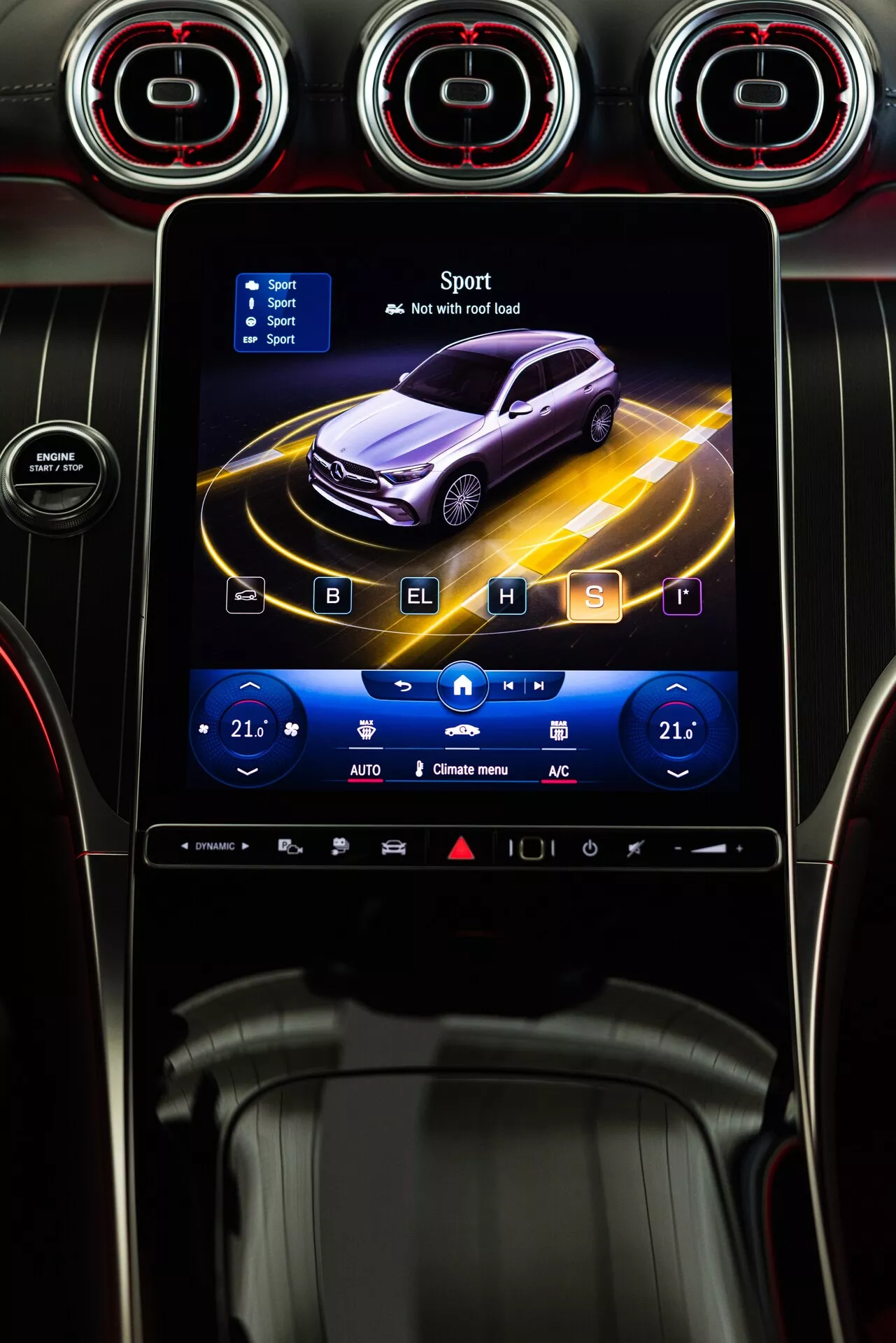 Console centrale du Mercedes GLC 2022 avec le système d'infodivertissement affichant une vue de profil animée d'une voiture violette foncée entourée de lignes jaunes dynamiques, en mode 'Sport' avec une icône de drapeau à damier. L'écran tactile est encadré par des aérateurs circulaires avec une finition métallique et des accents rouges. En dessous, les commandes tactiles de climatisation affichent des températures duales réglées à 21°C, avec des icônes pour le chauffage des sièges, la climatisation et le dégivrage. Panneau noir brillant avec des touches de contrôle tactiles supplémentaires en dessous. Le style indique une technologie avancée et une esthétique sportive.