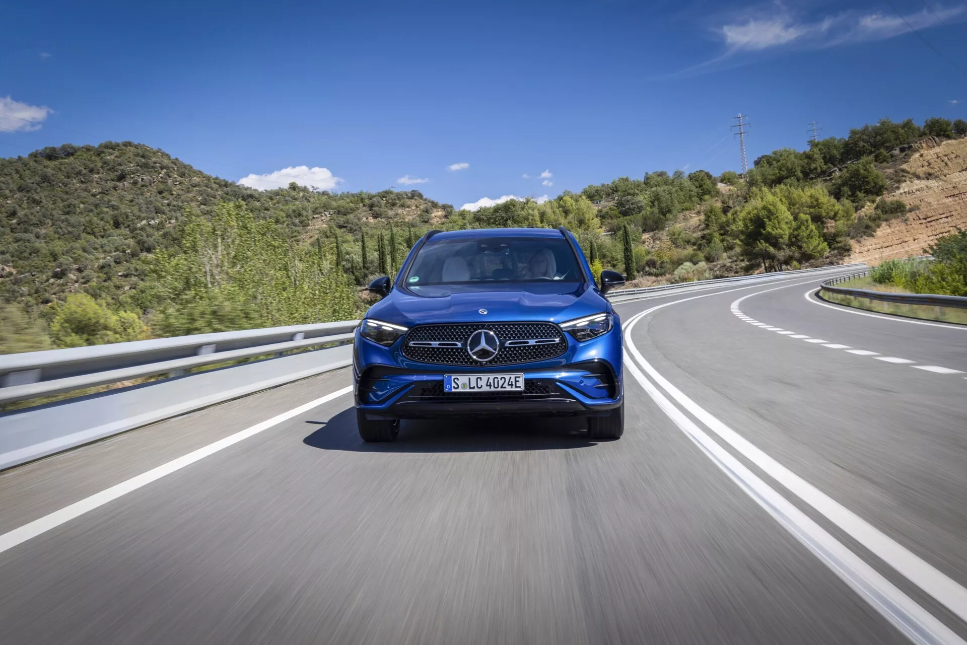 Vue frontale en angle bas d'une Mercedes GLC 2022 moderne roulant sur une autoroute ou une route rurale. Cette image met en évidence la calandre distinctive du véhicule, le design des phares, le style du pare-chocs avant, les grandes roues en alliage et les éventuels systèmes avancés d'assistance à la conduite (ADAS). L'éclairage est lumineux et naturel avec un fond de ciel bleu et de collines vertes, suggérant un mouvement sur l'asphalte avec des marques de voie de division.