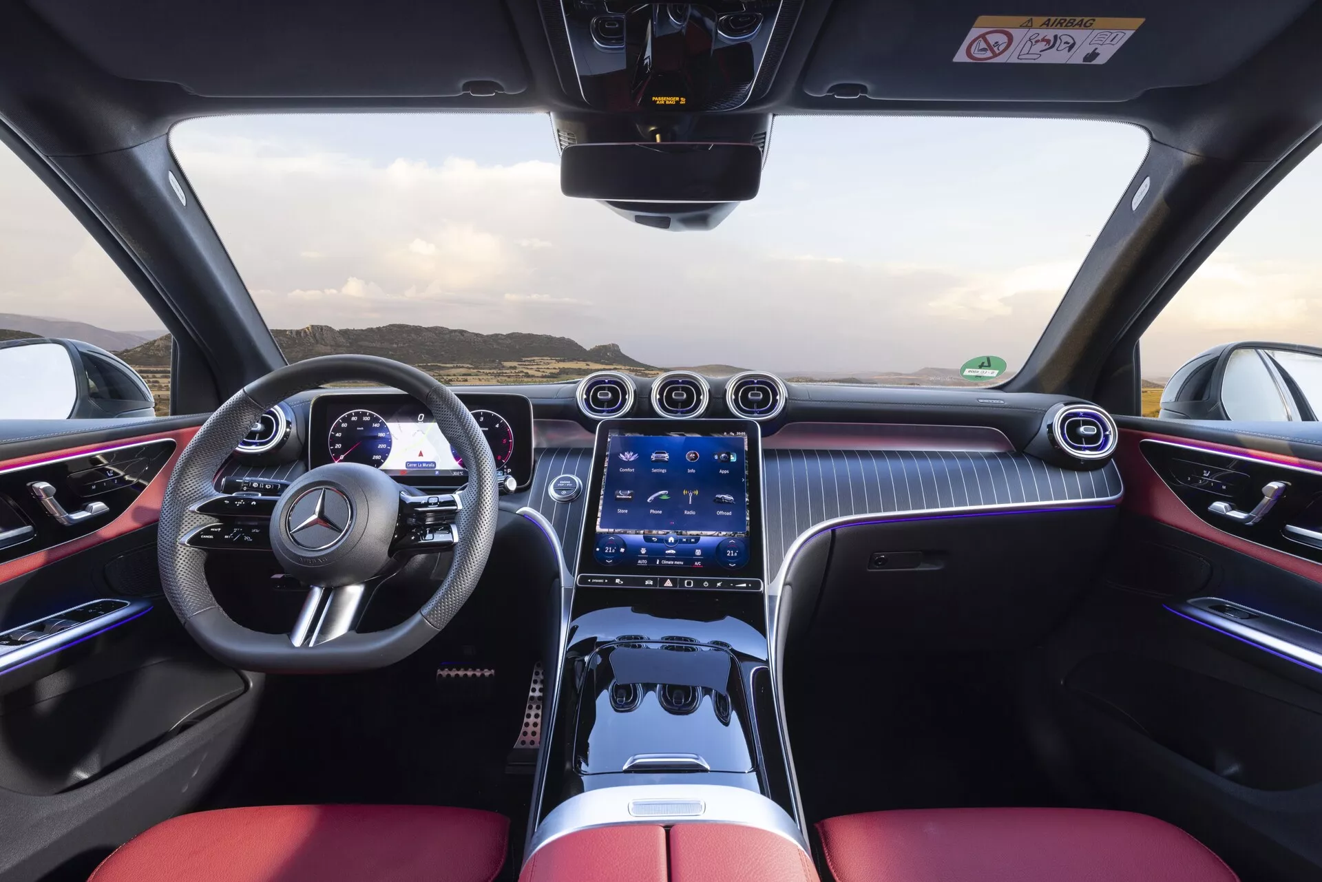 Vue intérieure de la Mercedes-Benz GLC 254 de 2022 depuis la perspective du conducteur, montrant le volant sport avec fonctions multiples, cockpit bicolore avec sièges de luxe noirs et rouges vifs, tableau de bord numérique, large système d'info-divertissement tactile, aérateurs chromés, boutons de commande climatiques physiques, motifs semblables à la fibre de carbone, éclairage d'ambiance, contrôle par pavé tactile et un design axé sur la haute performance.