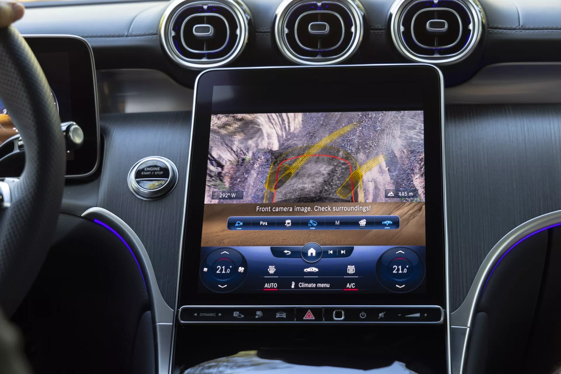 Photo en gros plan de l'intérieur d'une Mercedes GLC 2022 montrant la console centrale depuis la perspective d'un passager. L'accent est mis sur l'écran tactile verticalement orienté affichant la vue de la caméra frontale avec des lignes directrices de stationnement, les boutons de contrôle du climat avec des réglages de température à double zone de 21°C, des bouches d'aération circulaires au-dessus de l'écran et le bouton de démarrage/arrêt du moteur. L'image met en avant les systèmes d'infodivertissement avancés, l'aide au stationnement, le contrôle du climat à double zone et les matériaux intérieurs haut de gamme.