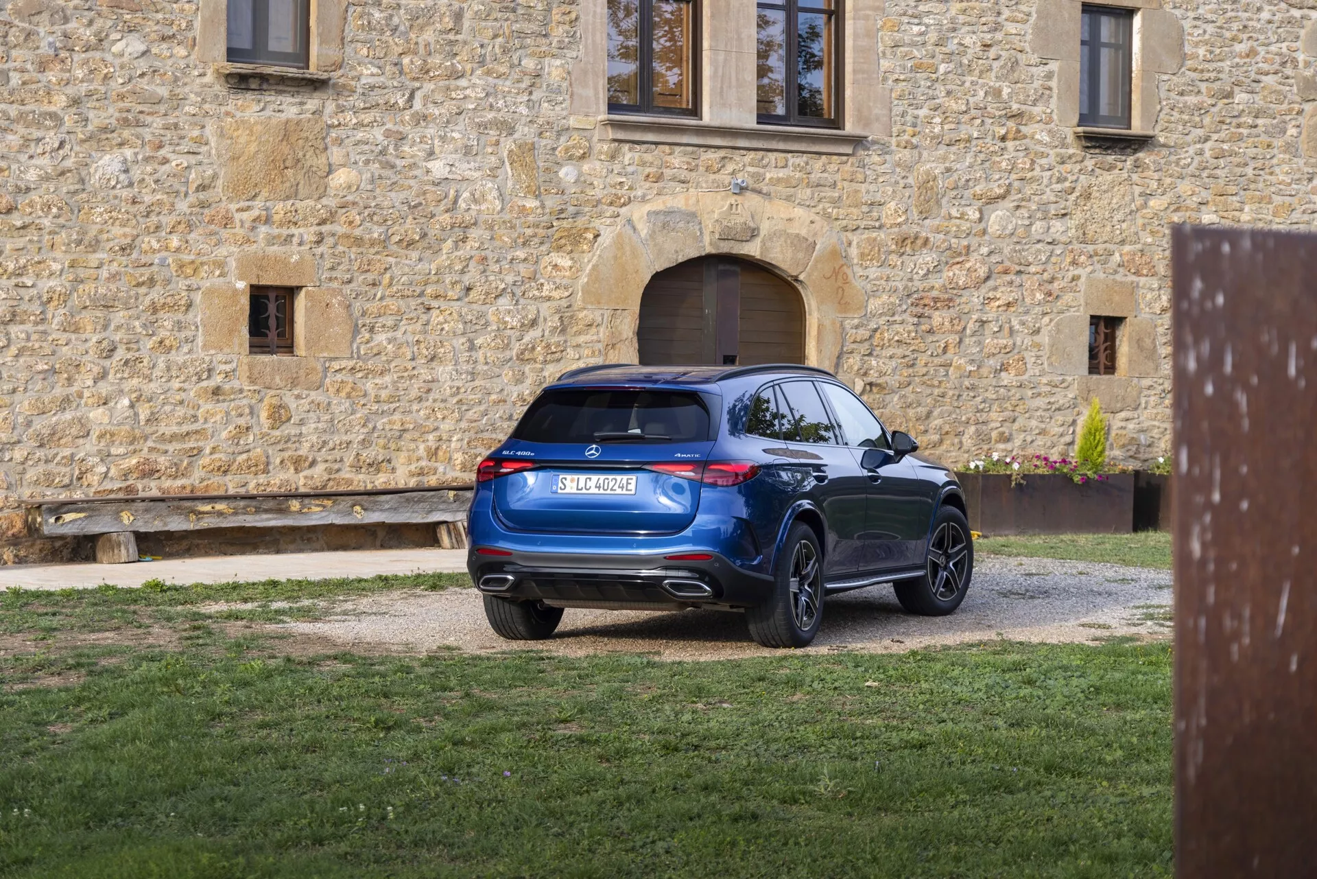 SUV moderne garé sur une allée de gravier vu de trois quarts arrière. Les feux arrière intègrent une technologie LED contemporaine harmonieusement intégrée au design. La lunette arrière légèrement inclinée vers l'avant contribue à un profil aérodynamique, complété par un becquet de toit. Le pare-chocs arrière comporte des capteurs ou des réflecteurs intégrés, suggérant la présence d'un système d'assistance au stationnement et présente des doubles embouts d'échappement intégrés dans la partie inférieure du pare-chocs. Le SUV arbore de grandes roues en alliage à plusieurs rayons et une surface bleue riche et propre, reflétant un aspect bien entretenu sur fond de bâtiment en pierre rustique.