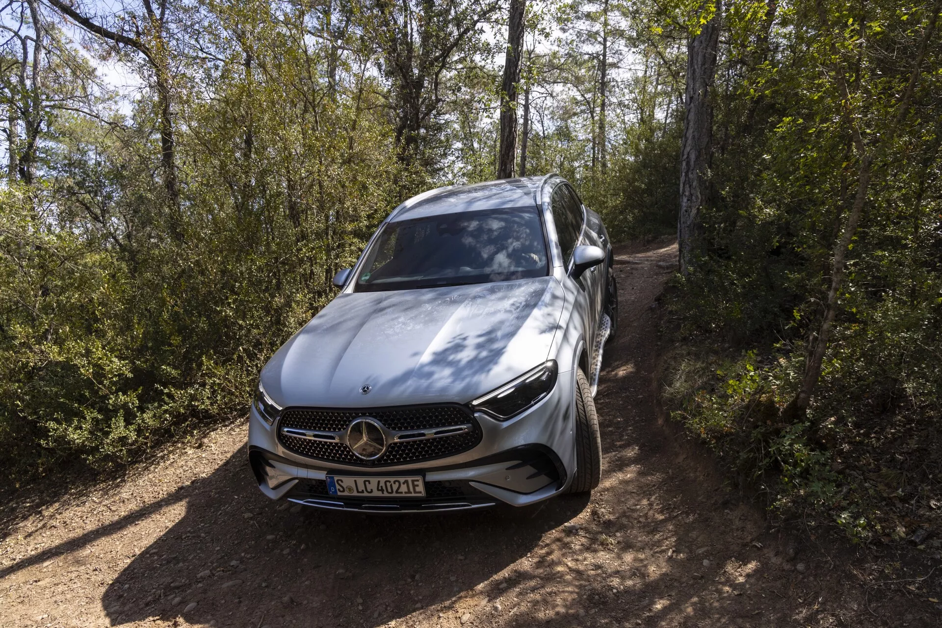 Voiture de luxe moderne, Mercedes-Benz GLC 2022, perspective de haut en avant montrant la calandre avant, le pare-chocs, le capot, le côté gauche y compris la porte du conducteur et le rétroviseur latéral, design contemporain élégant, emblème emblématique à trois branches de Mercedes-Benz, phares LED détaillés, roues en alliage à plusieurs rayons, badge potentiel EQ indiquant modèle électrique ou hybride, caractéristiques probablement ADAS, situé sur un chemin de terre entouré de végétation, peinture métallique argentée ou gris clair soulignant la qualité premium.