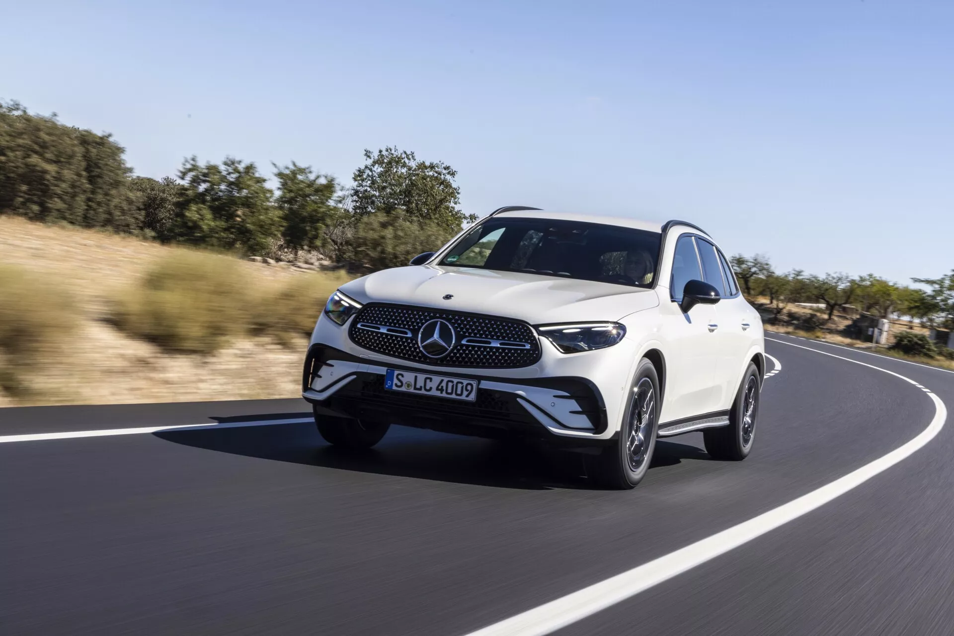 SUV crossover de luxe Mercedes GLC 2022 blanc roulant sur une route lisse en asphalte, décor naturel en arrière-plan sous un ciel dégagé. Vue dynamique en contre-plongée avec angle de trois quarts avant montrant les phares LED, la grille avant avec emblème, le design aérodynamique et les roues en alliage, soulignant la sensation de vitesse et de mouvement avec un léger flou de l'arrière-plan.