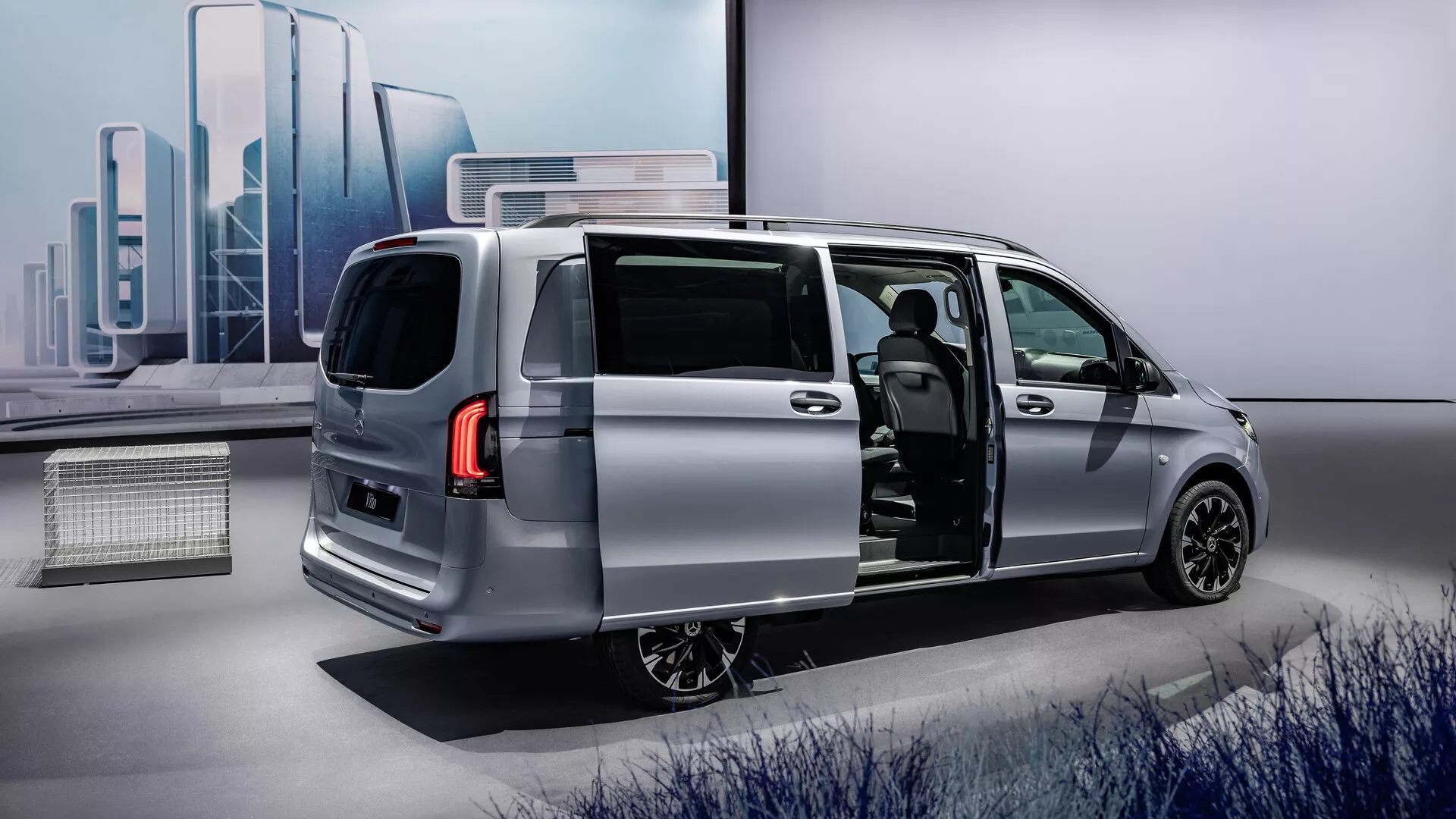 Monospace argenté Mercedes-Benz Vito 2024 (447 facelift) avec porte latérale coulissante ouverte montrant le design moderne élégant depuis un angle arrière trois-quarts, soulignant le profil aérodynamique du véhicule, les feux arrière LED, les roues en alliage et un aperçu de l'intérieur avancé avec un matériau de siège potentiellement premium et un tableau de bord moderne.