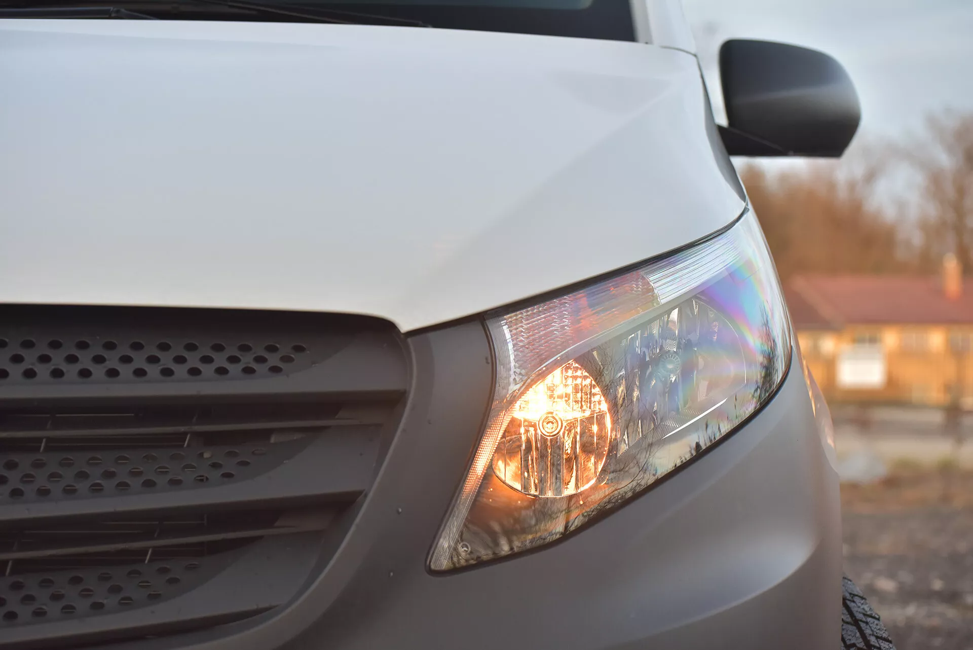 Vue de côté en gros plan d'un Mercedes-Benz Vito Tourer blanc de 2014, se concentrant sur le phare allumé, une partie de la grille et une section du capot, mettant en lumière ses phares halogènes et son design aérodynamique sous une lumière douce.