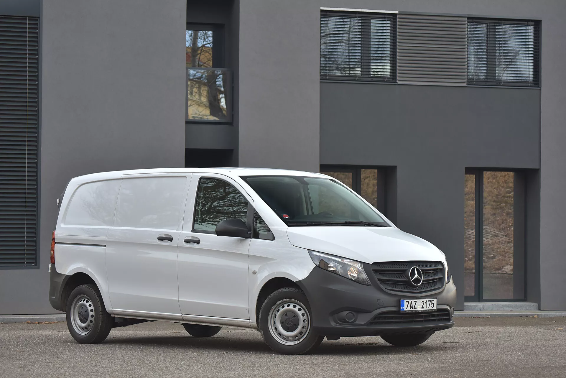 Fourgonnette Mercedes-Benz Vito Tourer de 2014 en blanc stationnée sur un tarmac, affichant un profil avant et latéral avec une vue claire de l'emblème de la grille et des phares modernes, prise sous une lumière diurne nuageuse devant un bâtiment minimaliste
