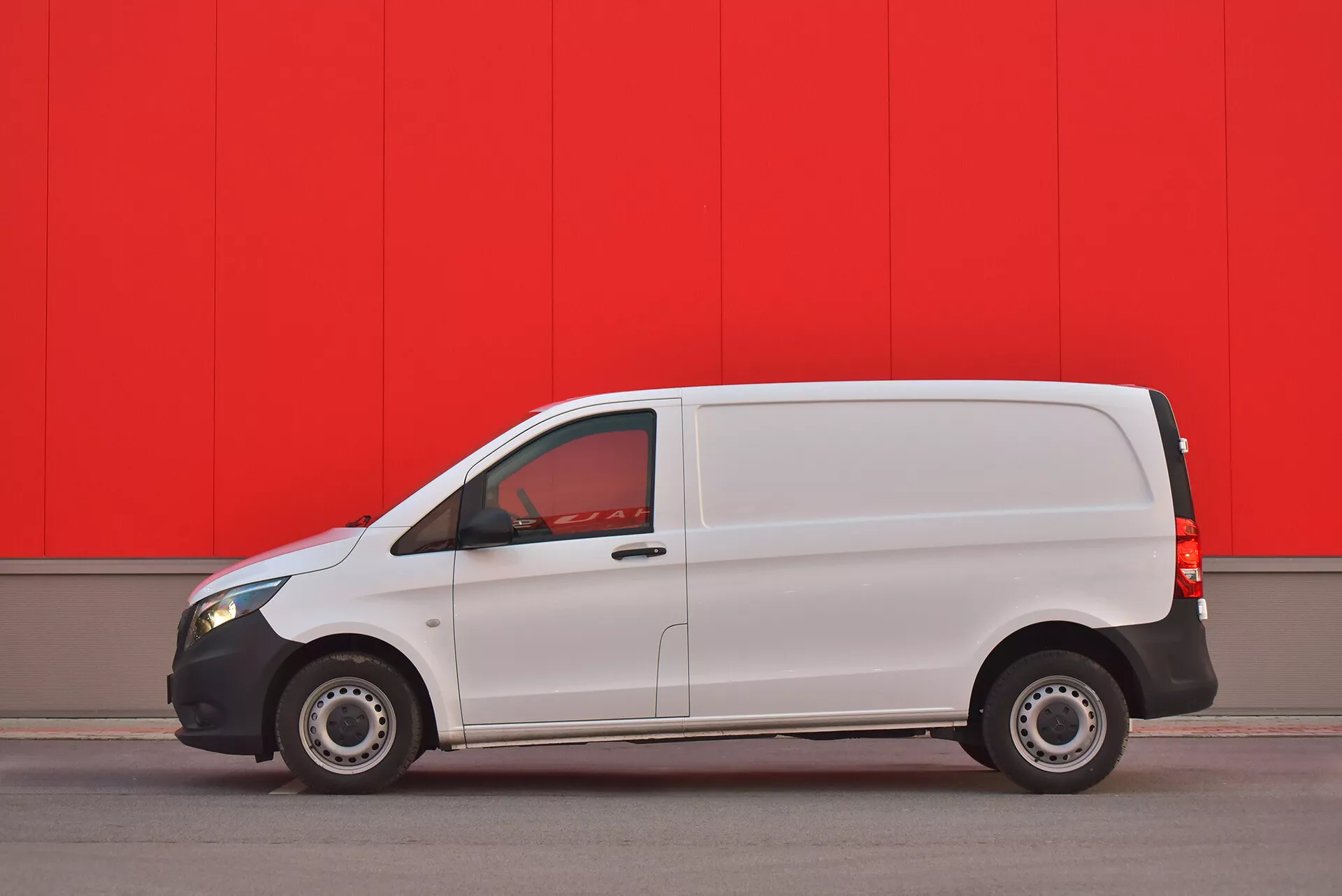 Fourgon commercial blanc Mercedes-Benz Vito Tourer de 2014 garé parallèlement contre un mur rouge vibrant, montrant le profil latéral avec porte visible, fenêtres et roues en acier capturées depuis un angle bas pour souligner la conception et les caractéristiques du véhicule