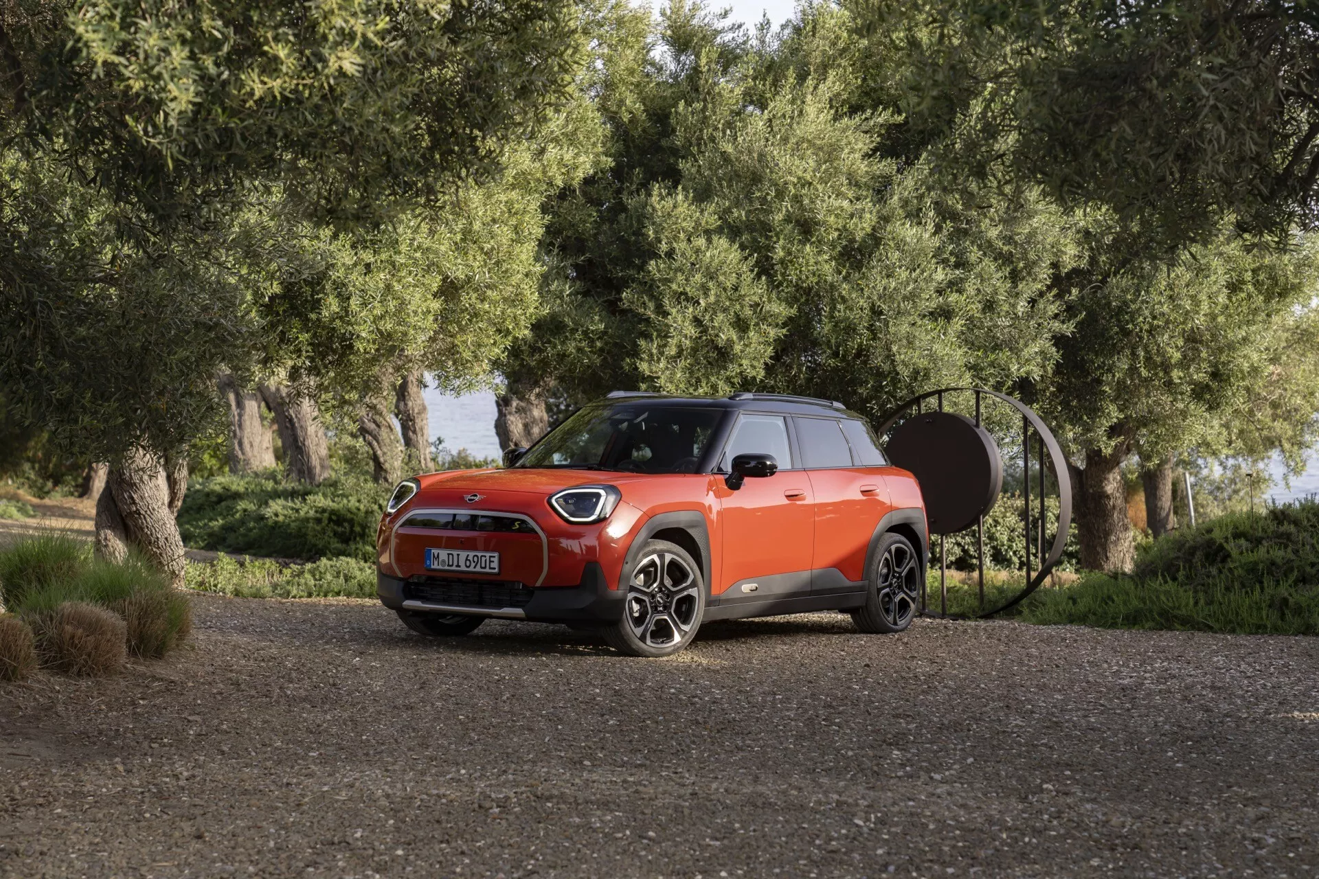 Mini Aceman 2024 (J05) vue de trois quarts avant sur un chemin de gravier avec verdure, présentant ses phares LED, jantes en alliage à multiples branches, couleur de carrosserie bicolore avec toit noir, design sportif du capot et silhouette compacte de SUV