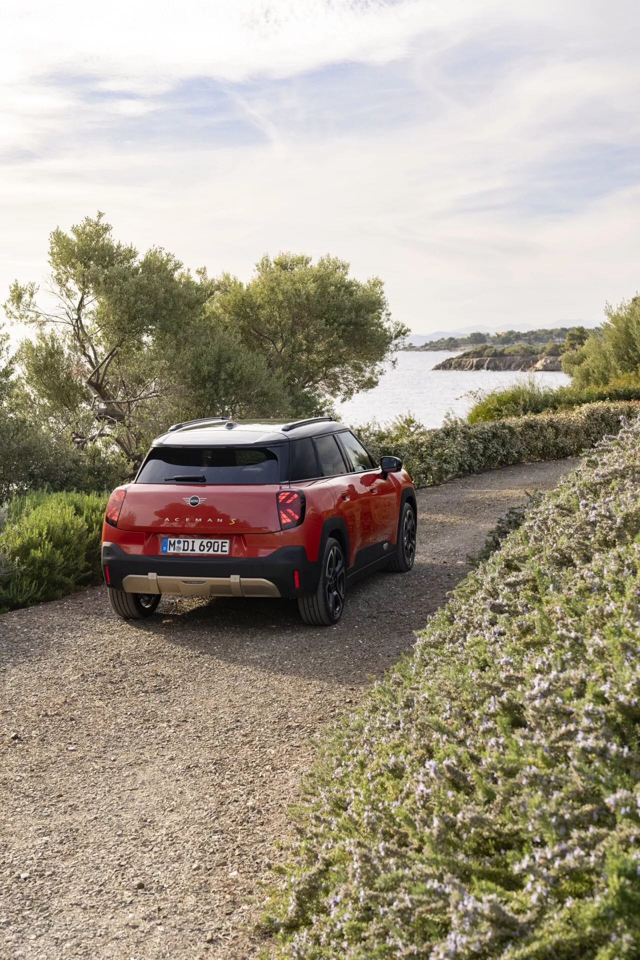 SUV compact Mini Aceman 2024 en rouge, montrant vue arrière trois quarts près d'un lac ou mer calme, soulignant les feux arrière et le contour, entouré d'un paysage naturel sur un chemin de gravier, indiquant des capacités tout-terrain et des caractéristiques de design extérieur