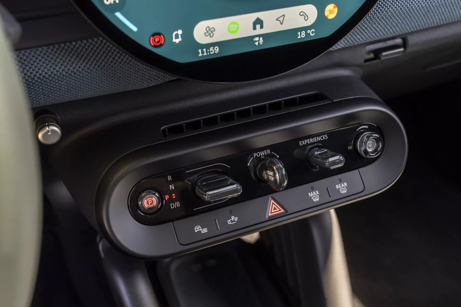 Vue rapprochée des commandes de climatisation et du sélecteur de mode de conduite du Mini Aceman 2024 (JO5), mettant en vedette les réglages de climatisation bi-zone, les boutons de circulation d'air, le commutateur de feux de détresse et le cadran rotatif de mode de conduite avec un aperçu de l'affichage numérique indiquant l'heure et la température extérieure