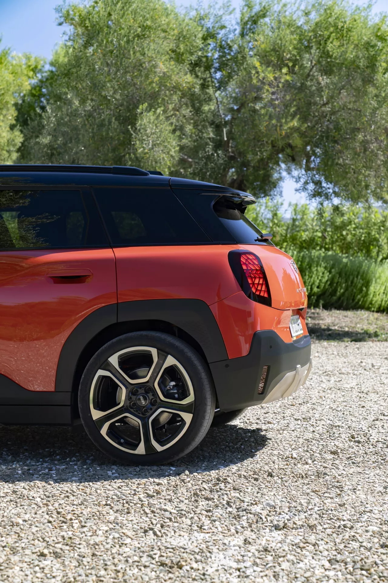 Vue arrière trois quarts du Mini Aceman (J05) 2024 montrant son côté droit sous un angle surélevé, présentant une finition mate rouge-orange vibrante, des jantes en alliage à cinq doubles rayons, une configuration de feux arrière à LED, un pare-chocs arrière avec capteurs de stationnement et un essuie-glace arrière, dans un cadre extérieur verdoyant.