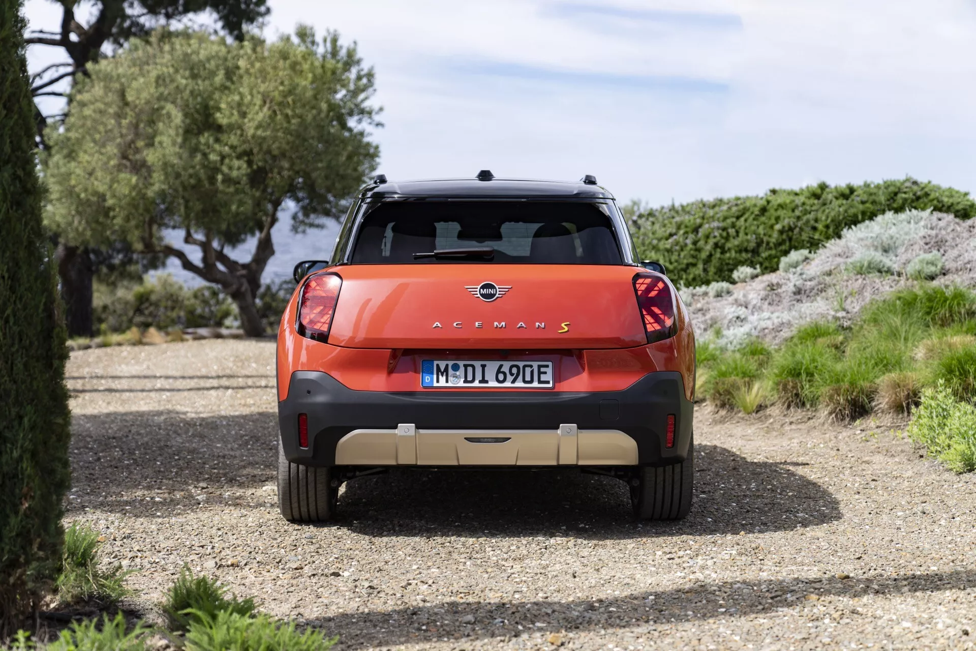 Vue arrière du Mini Aceman 2024 (JO5) garé sur un chemin de gravier présentant emblème, nom du modèle avec désignation 'E', pare-chocs arrière contrastant, feux arrière LED et barres de toit entourés par un paysage naturel