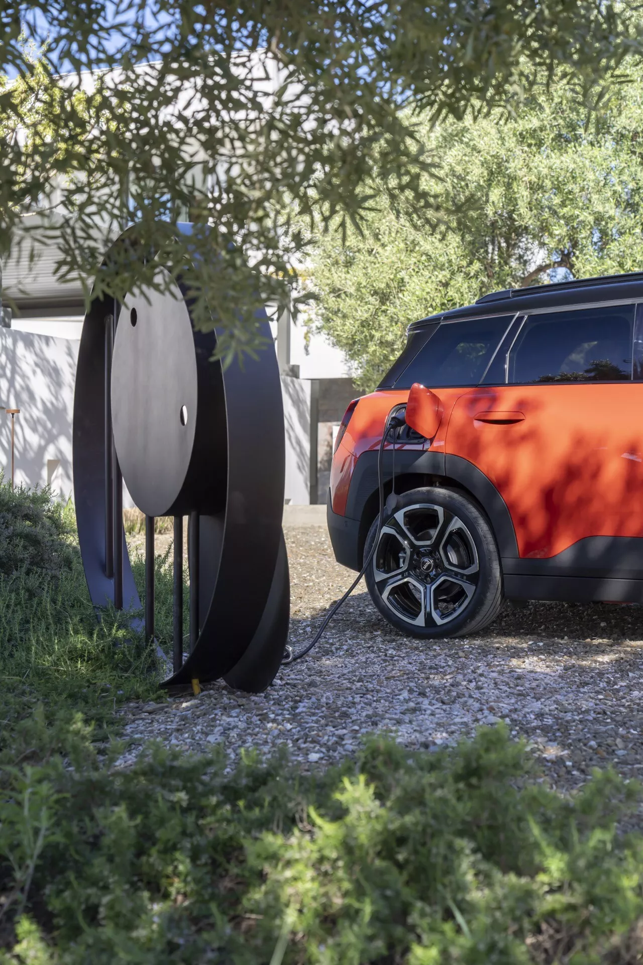 Vue partielle d'une Mini Aceman 2024 (JO5) montrant le côté arrière droit avec une roue en alliage à multi-rayons, une carrosserie vibrante et un design moderne de feu arrière, encadrée par du feuillage naturel et un élément décoratif extérieur circulaire, sous la lumière naturelle du jour