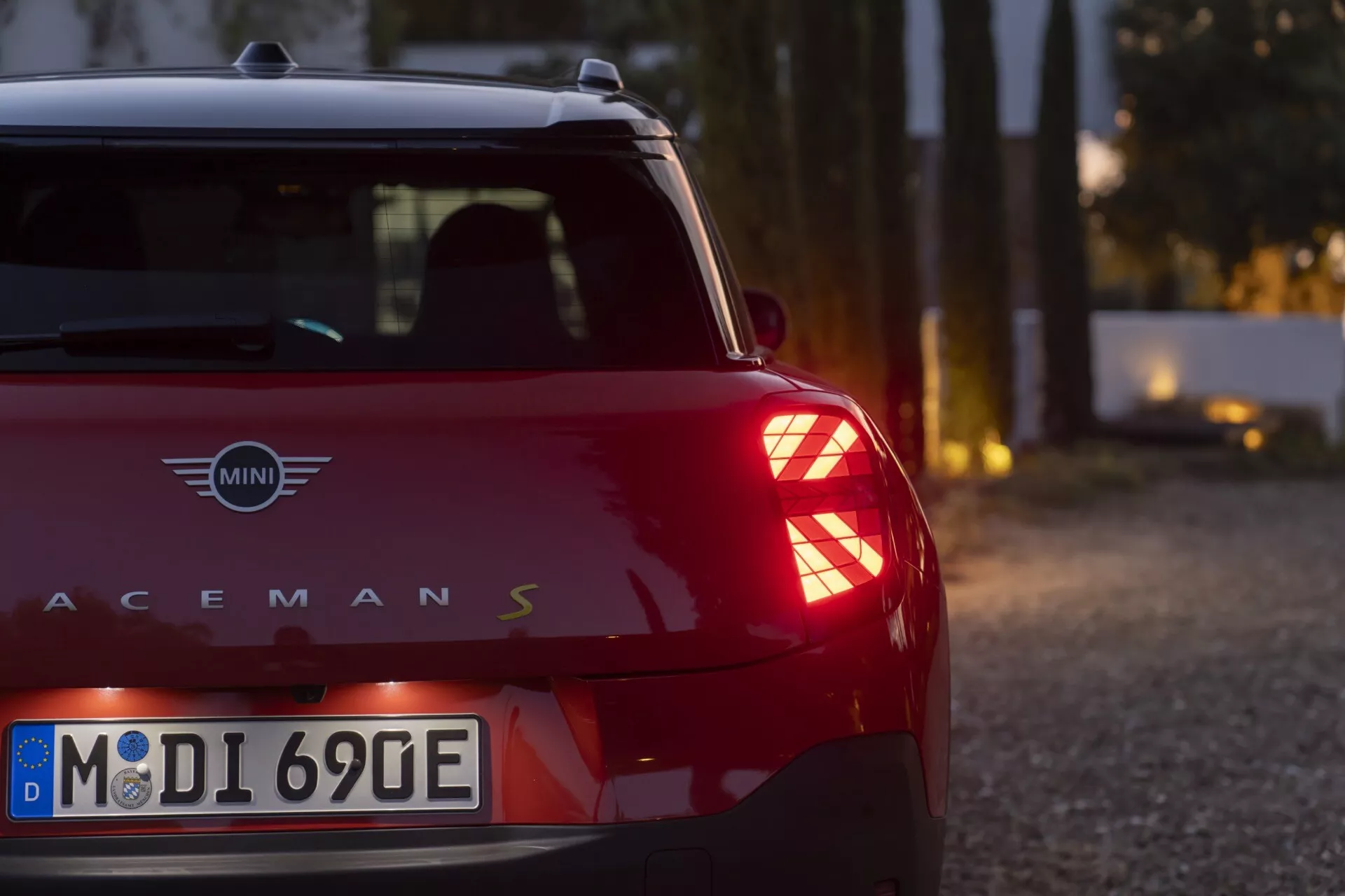 MINI Aceman S 2024 vue d'une perspective arrière basse trois-quarts mettant en lumière ses feux arrière LED uniques avec motif Union Jack, carrosserie rouge foncé avec toit noir contrastant, et plaque d'immatriculation européenne écologique, dans un cadre serein au crépuscule