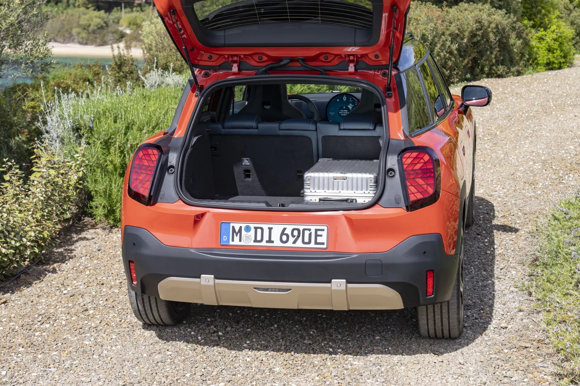 Vue arrière d'un SUV compact Mini Aceman (J05) orange de 2024 garé sur un gravier, avec le hayon ouvert exposant la zone de chargement, équipé de capteurs de stationnement, feux arrière uniques, tapis de coffre aftermarket, badge de modèle '690E', et essuie-glace arrière, sur fond de paysage naturel