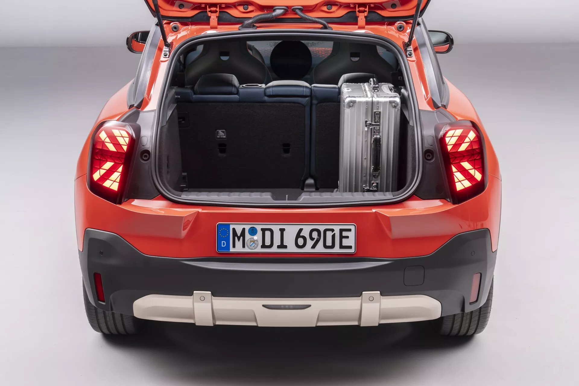 Vue arrière du MINI Aceman (JO5) 2024 montrant le coffre spacieux avec bagage métallique, feux arrière LED complexes, capteurs de stationnement sur le pare-chocs et finition couleur orange-rouge éclatante