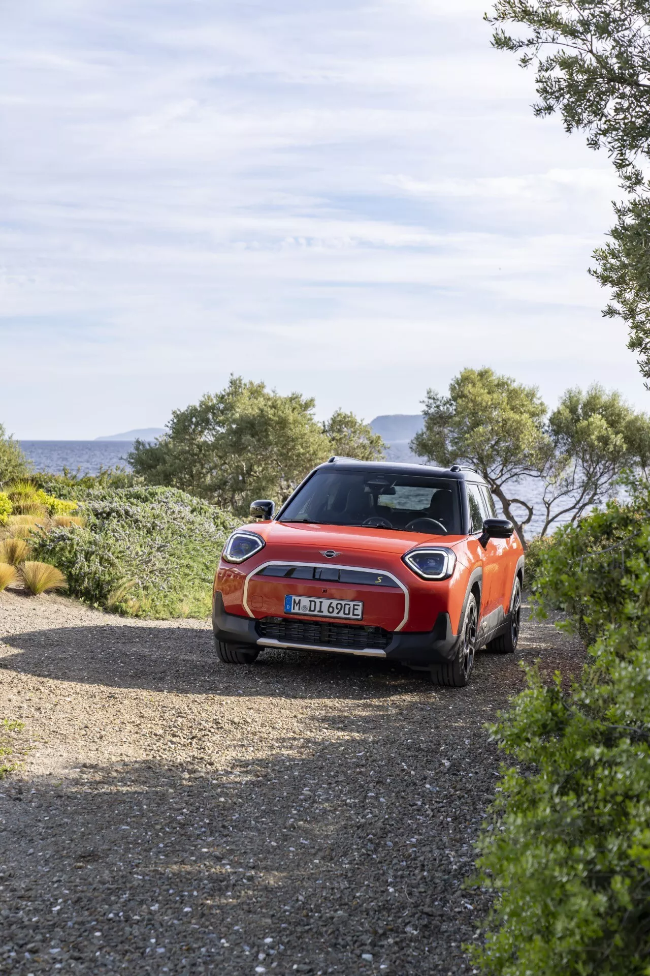 Mini Aceman 2024 (J05) garée sur un chemin de gravier, montrant des feux arrière LED, une finition brillante orange vibrante, des roues en alliage, une vitre arrière teintée, des rails de toit, une antenne aileron de requin, un style de carrosserie hatchback, entourée d'un décor naturel avec des capacités potentielles de conduite hors route, sous la lumière naturelle du jour