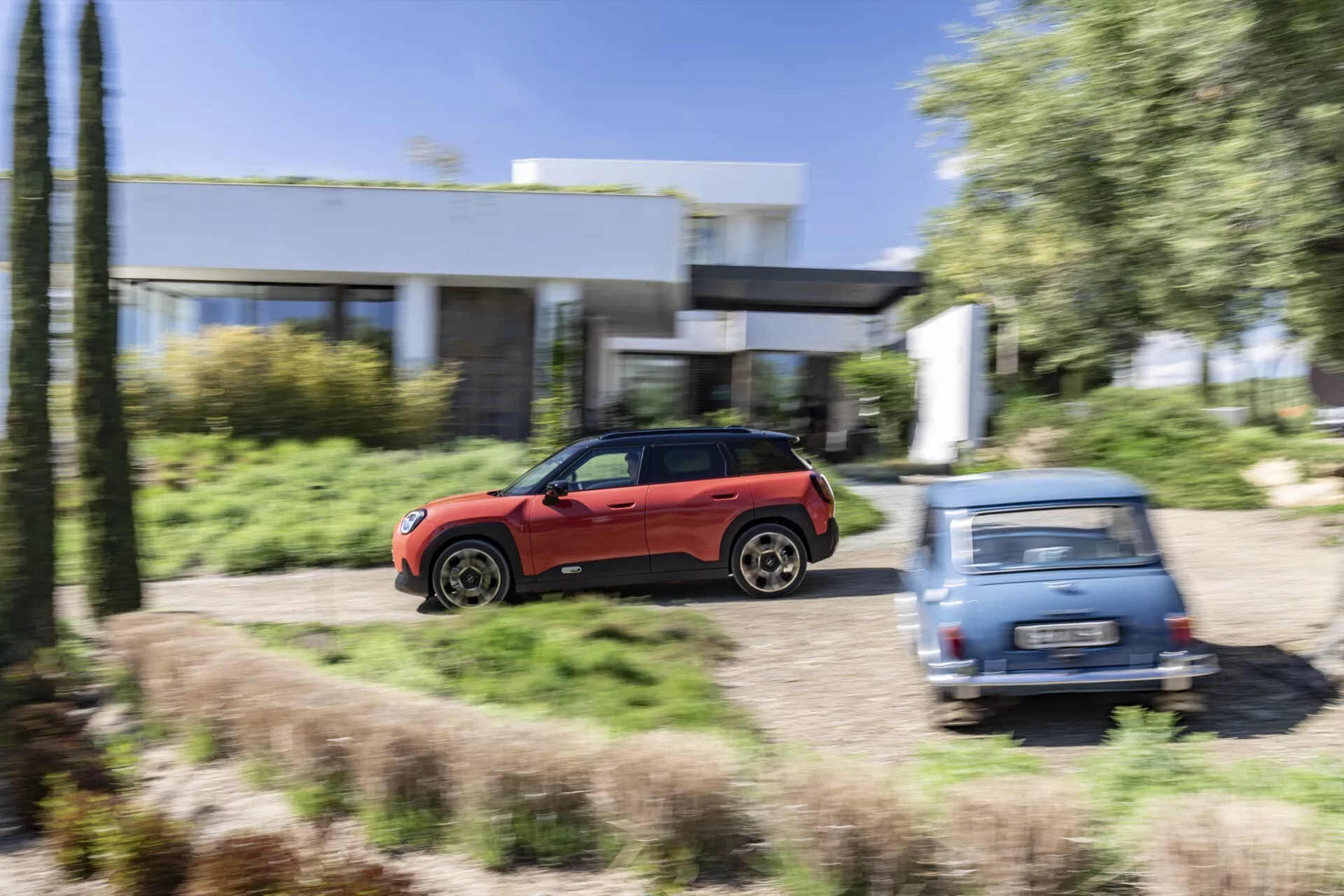 Mini Aceman rouge 2024 (J05) en mouvement capturé de profil, mettant en évidence son design aérodynamique élégant et ses grandes roues en alliage, suggérant une capacité tout-terrain, sur un fond flou de paysage résidentiel et verdoyant.