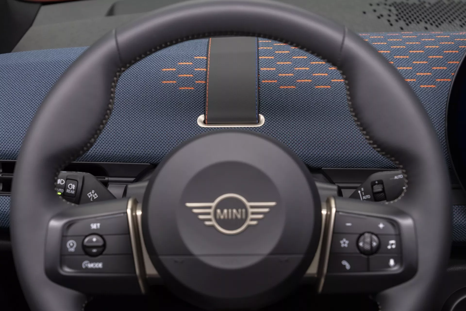 Vue rapprochée des trois quarts inférieurs du volant de MINI Aceman 2024 avec logo, boutons de contrôle pour les médias et le régulateur de vitesse, design sportif avec repose-pouces et vue partielle d'un tableau de bord numérique.