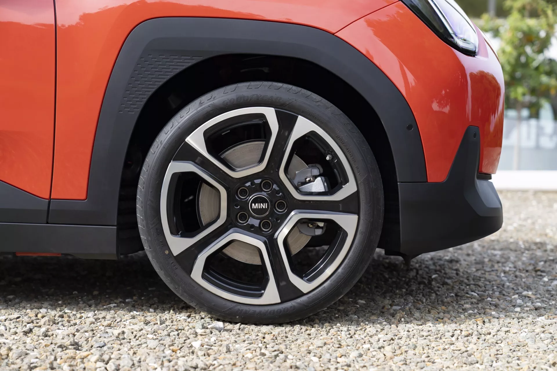 Vue rapprochée de la roue avant droite et du bas de l'aile du MINI Aceman 2024 en orange vif avec jante alliage à cinq branches bicolore et logo MINI, montrant le motif de la bande de roulement du pneu et la garniture noire de l'arche de roue sur une surface en gravier.