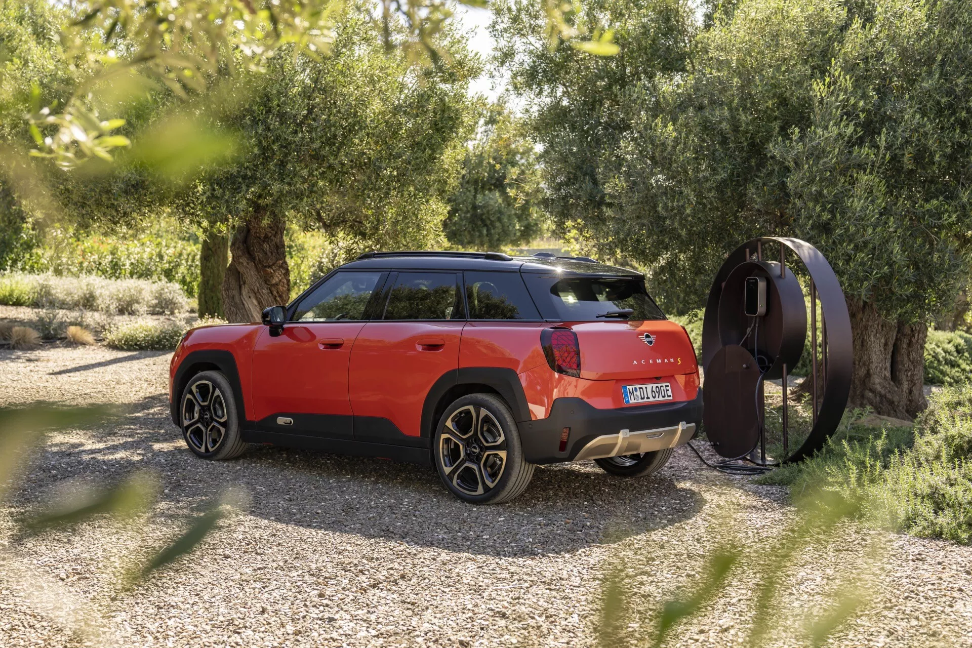 Mini Aceman 2024 (J05) en rouge vibrant montrant son profil latéral élégant et une vue partielle arrière avec des éléments de conception distinctifs, garée à l'extérieur parmi la verdure avec une station de charge à proximité, soulignant son potentiel en tant que SUV électrique ou hybride rechargeable.