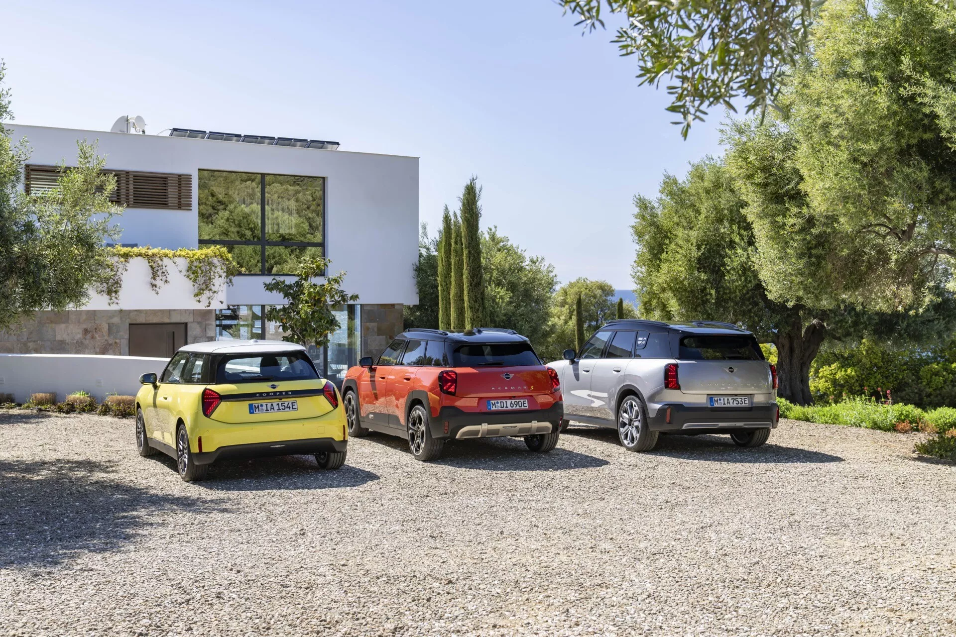 Modèles Mini Aceman (J05) 2024 en jaune vif, rouge orangé et argent garés sur gravier devant un bâtiment résidentiel moderne, présentant des vues arrière trois quarts avec un focus sur les éléments de design contemporain tels que les lignes aérodynamiques et l'éclairage moderne