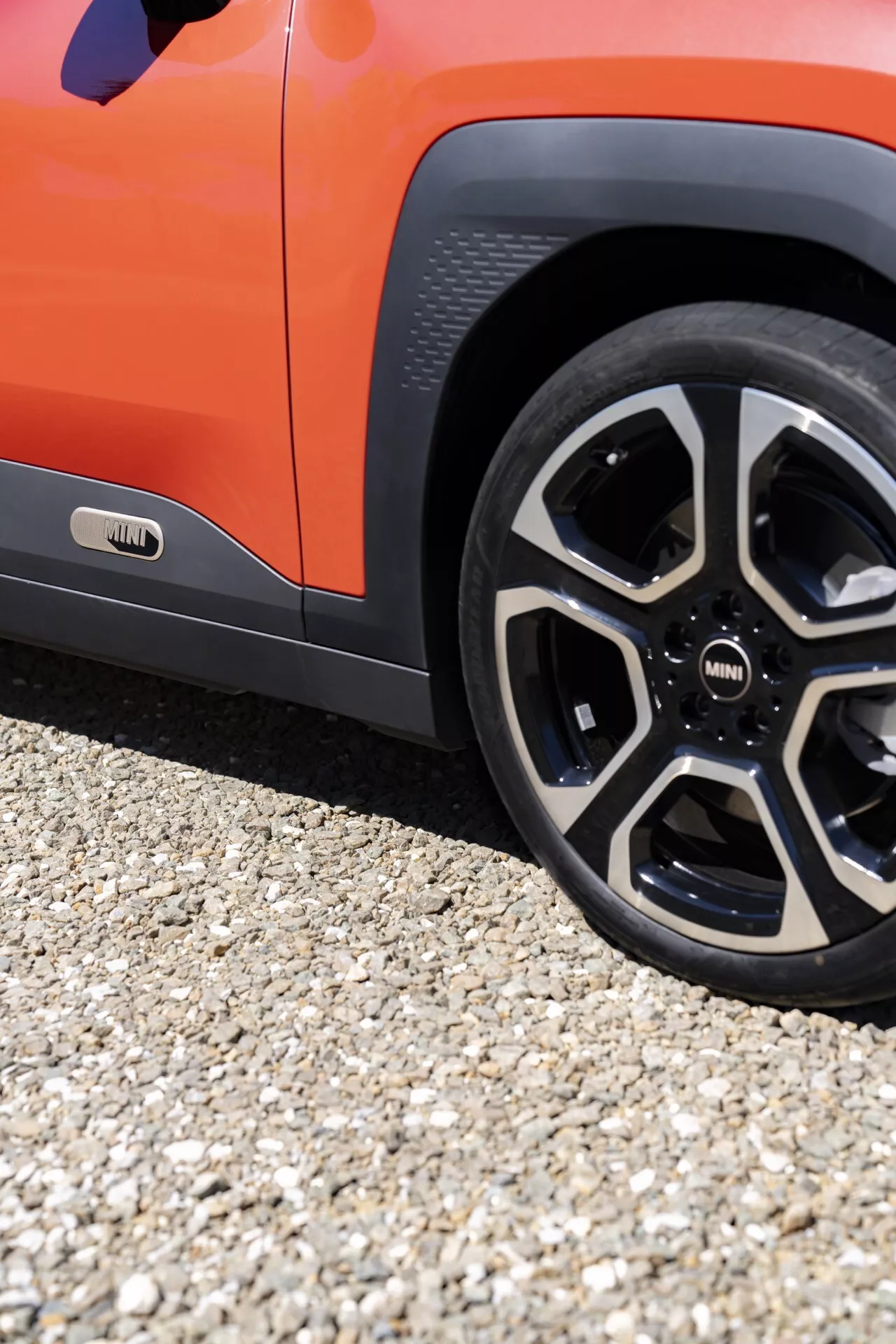 Vue détaillée du profil latéral inférieur du Mini Aceman 2024 (JO5), incluant un gros plan de la roue arrière avec un design moderne à multiples rayons et une partie de la porte avant orange, mettant en lumière des caractéristiques telles que le badge sur le seuil de porte, suggérant possiblement un modèle électrique ou hybride, garé sur une surface de gravier
