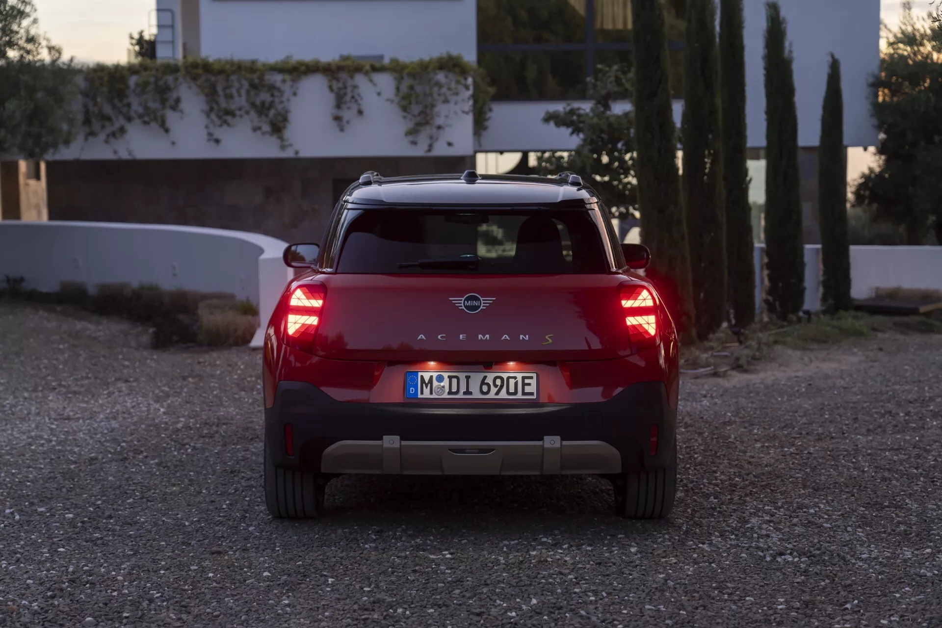 Vue arrière du SUV compact Mini Aceman (J05) de 2024 en rouge à l'aube ou au crépuscule avec les feux allumés, présentant des feux arrière LED distinctifs, un emblème de marque, une plaque d'immatriculation européenne, un spoiler arrière avec feu de freinage intégré, des capteurs de stationnement et des indications potentielles de véhicule électrique sans échappements visibles, sur fond résidentiel.