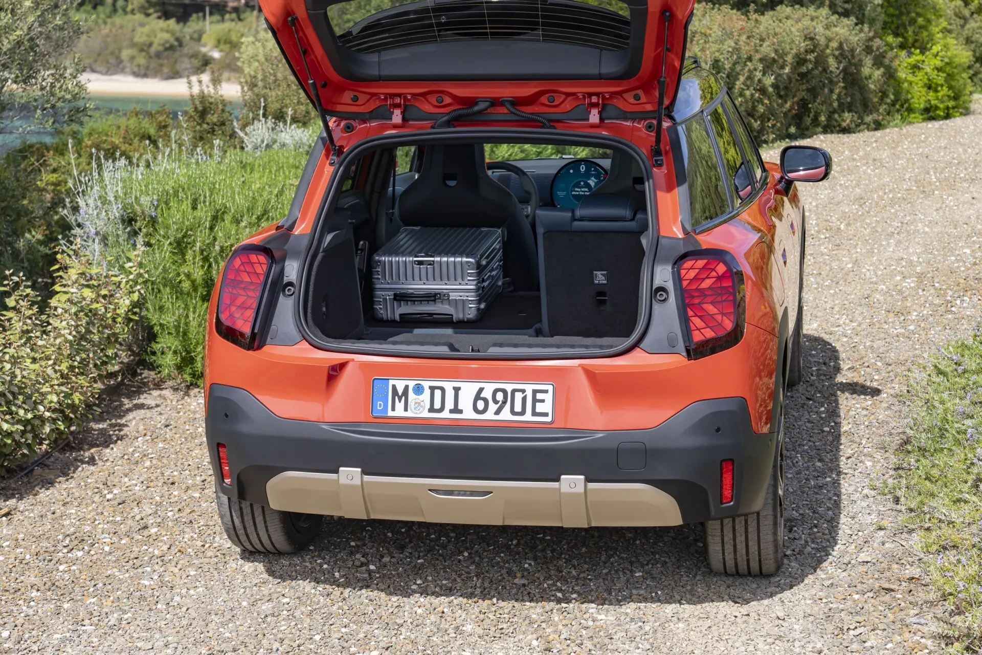Vue arrière de la hatchback orange Mini Aceman (J05) 2024 avec le coffre ouvert révélant un dispositif de charge VE, garée sur un gravier entouré de verdure