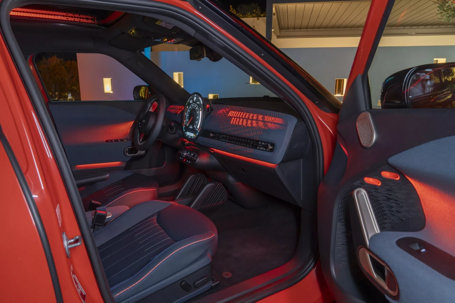 Vue intérieure de la Mini Aceman 2024 (J05) côté conducteur avec porte ouverte, montrant tableau de bord illuminé, écran d'infodivertissement, volant multifonction, sièges en tissu de haute qualité et caractéristiques modernes de l'habitacle