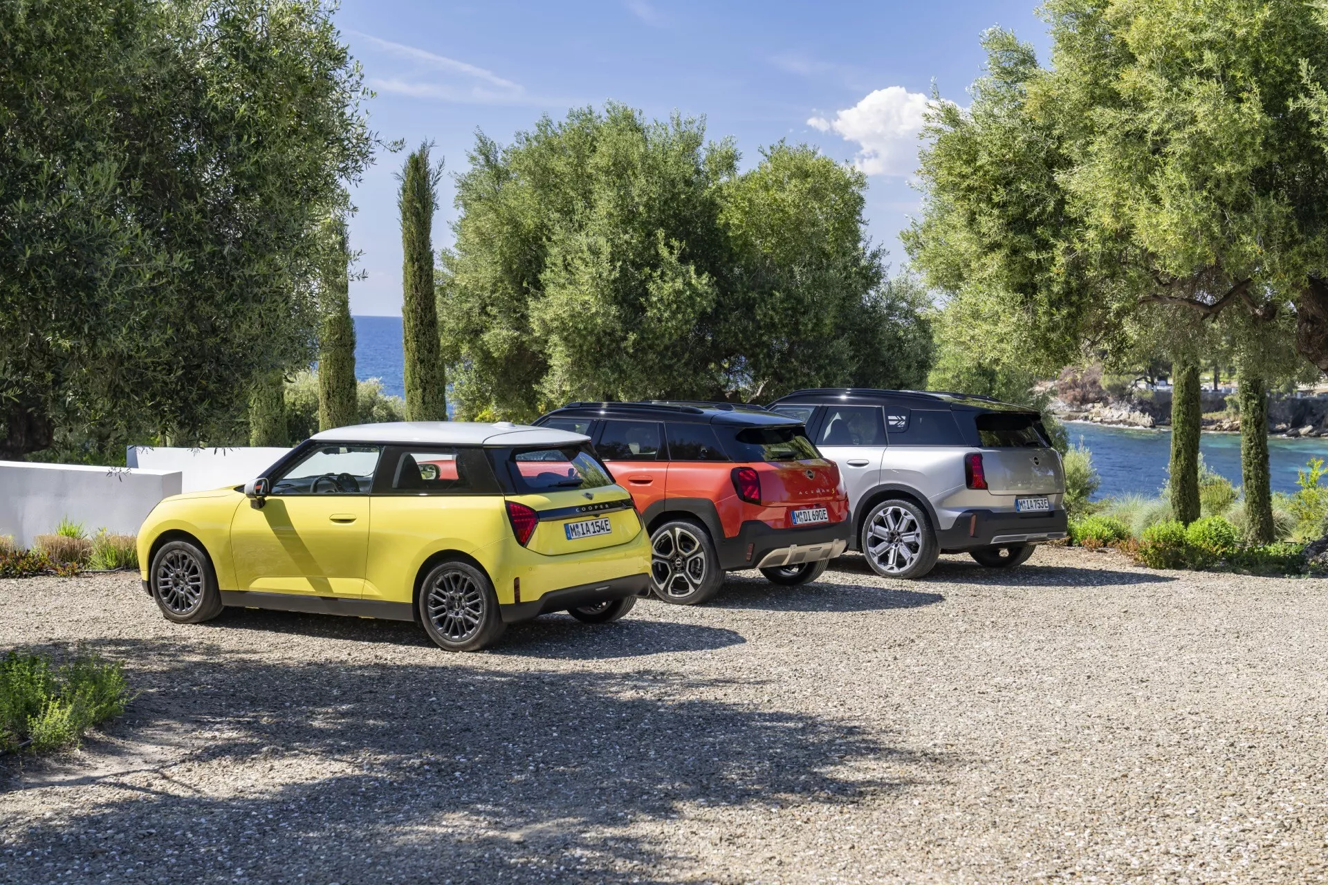 Mini Aceman 2024 (J05) en jaune vif avec un design de hayon moderne garé à côté d'un élégant SUV rouge et un sophistiqué crossover argent face à un paysage lacustre pittoresque avec une végétation luxuriante, encapsulant une atmosphère de vacances paisibles