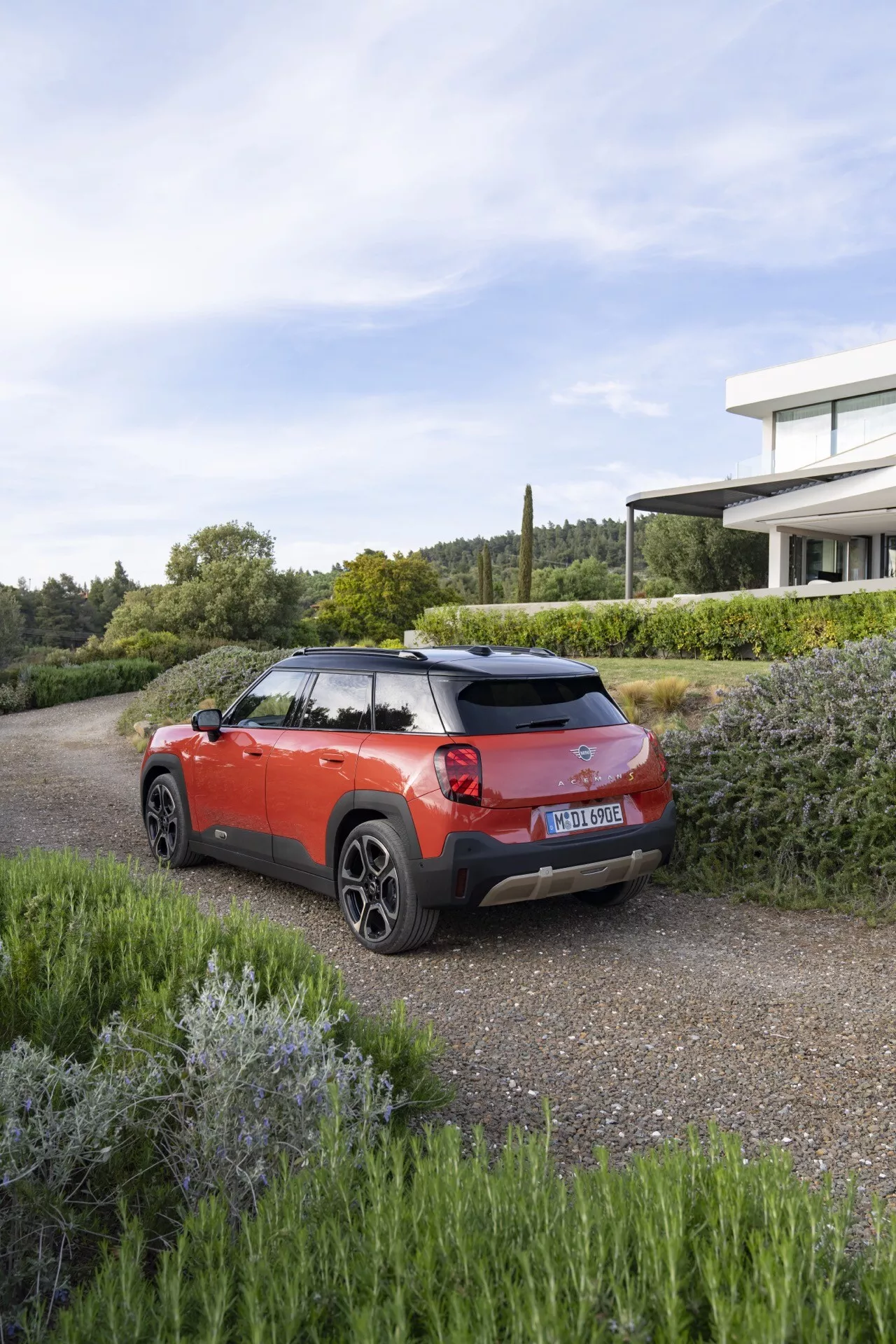 Mini Aceman 2024 (J05) en rouge avec des détails noirs, SUV moderne sur gravier avec verdure, présentant les points forts du design arrière et latéral sous une lumière du jour claire