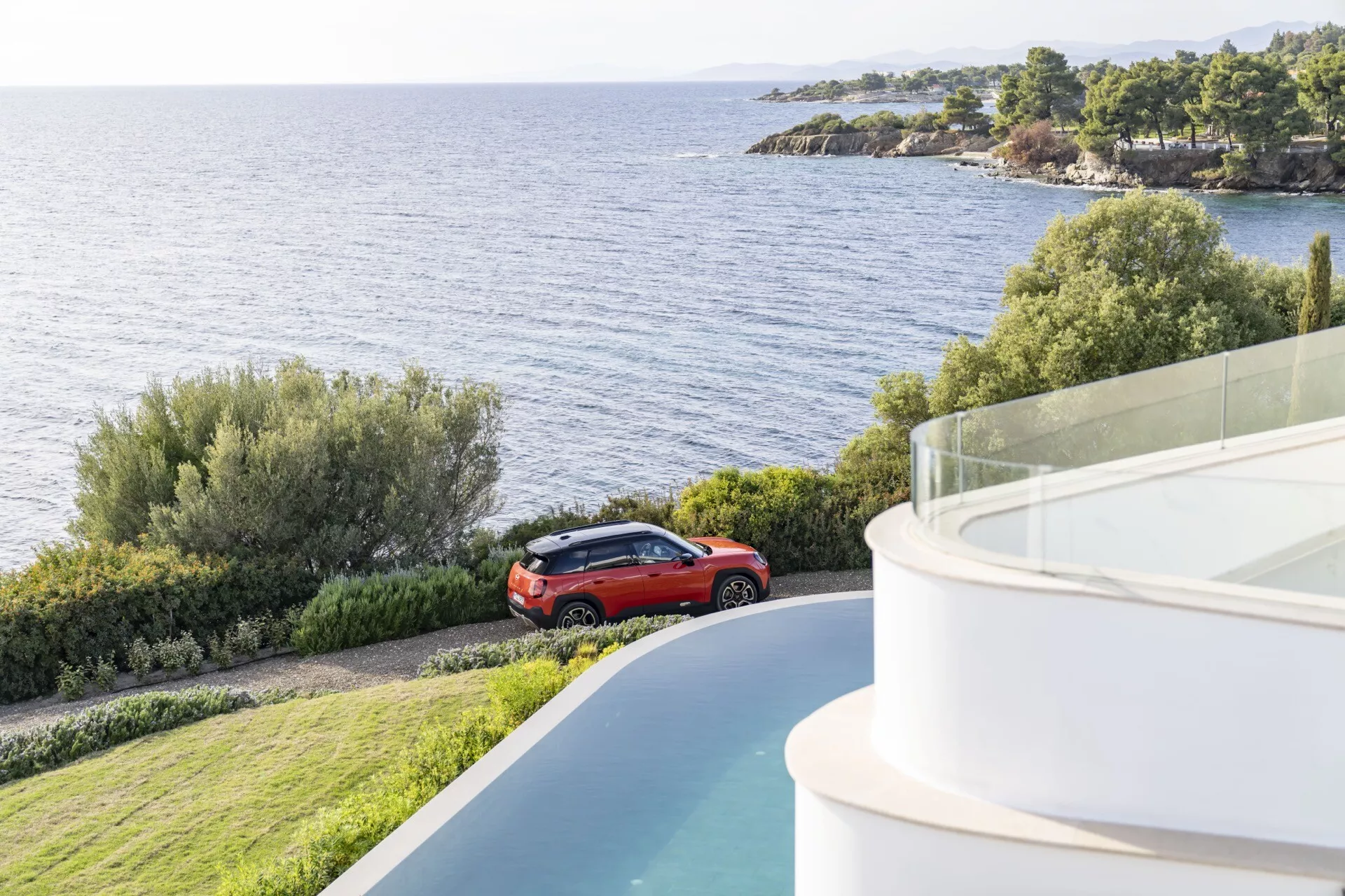 Mini Aceman (J05) de 2024 en rouge garée près d'une côte ou d'un lac pittoresque, vue sous un angle élevé montrant son design crossover avec des jantes en alliage et un bâtiment moderne aux détails architecturaux courbés au premier plan