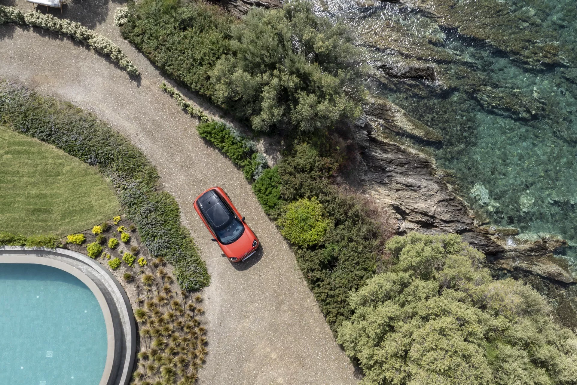 Mini Aceman 2024 (J05) en rouge vue de haut montrant son design compact hatchback, son toit ouvrant et ses rétroviseurs latéraux, positionnée sur un chemin courbé à côté de l'eau avec une végétation dense et une partie d'une piscine circulaire visible, soulignant sa connexion avec les loisirs et l'aventure en plein air.