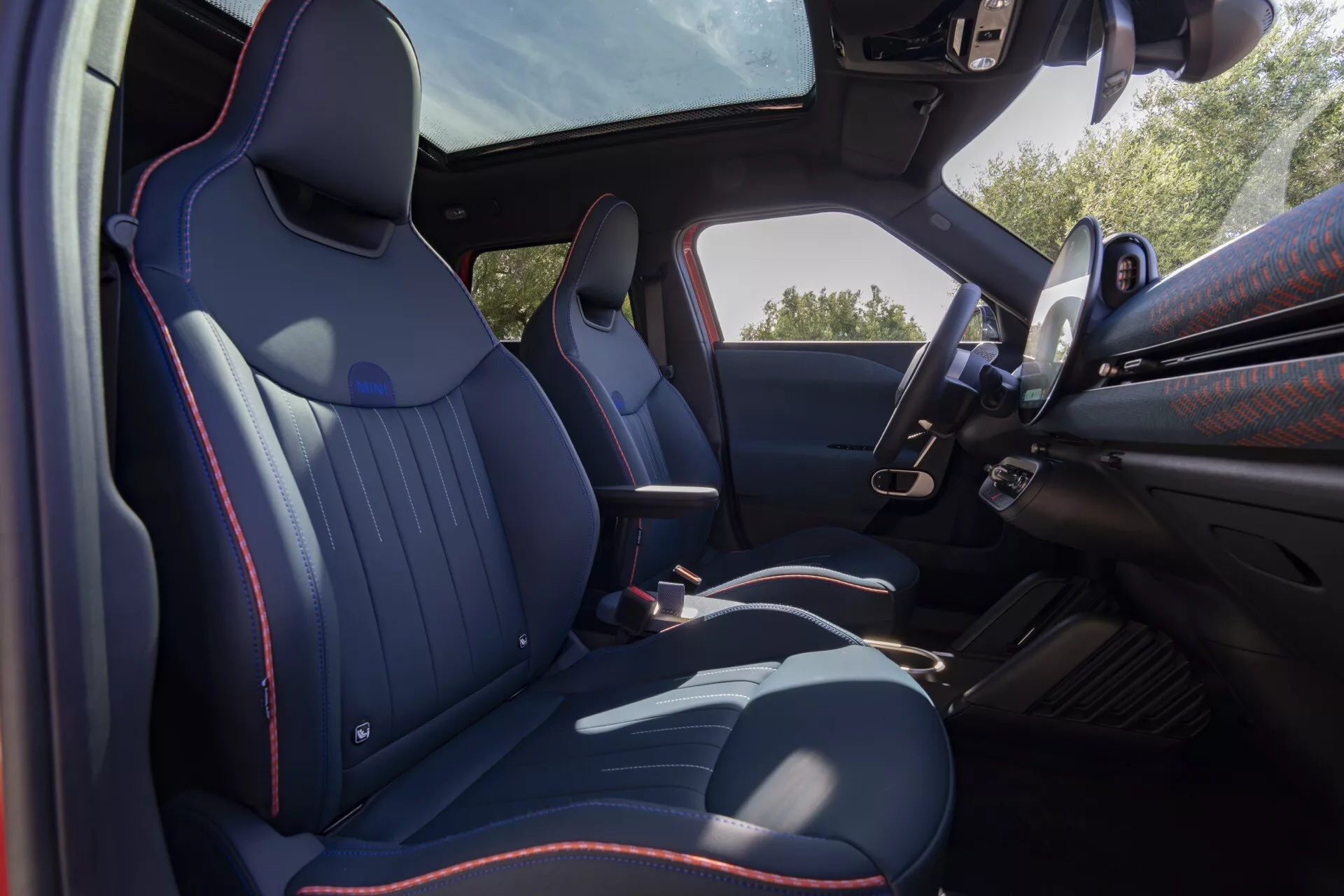 Vue intérieure du Mini Aceman 2024 (JO5) depuis l'arrière montrant des sièges avant profilés en cuir foncé et coutures rouges, panneau de porte du conducteur, volant multifonction, console centrale, toit panoramique et transmission automatique.