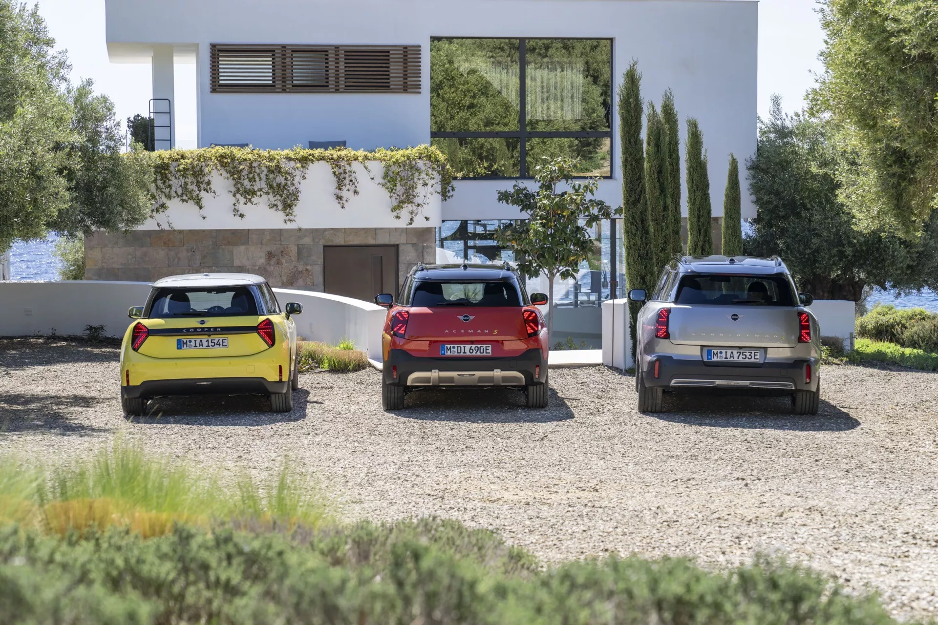 Trois hatchbacks et SUV Mini Aceman 2024 garés devant un bâtiment à l'architecture moderne sous un ciel dégagé, mettant en avant leur design arrière, feux arrière LED et couleurs distinctives jaune, rouge et argent.