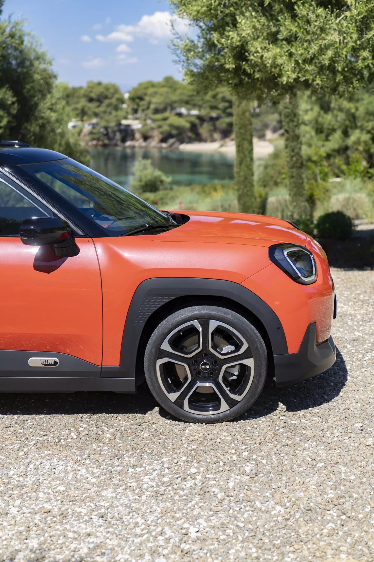 Vue en contre-plongée du quart avant latéral du Mini Aceman 2024 (J05) sur gravier, présentant une roue en alliage, un passage de roue noir et une carrosserie orange vif avec un lac naturel et des arbres en arrière-plan.