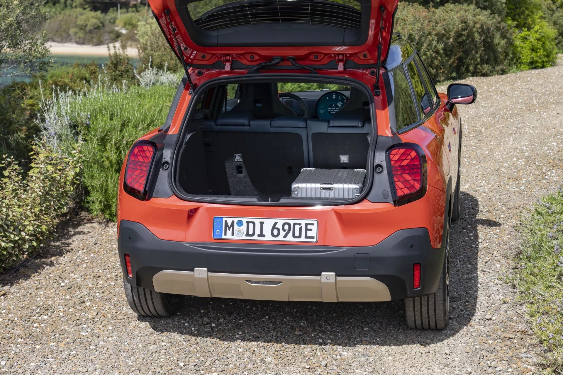 Vue arrière en plongée du Mini Aceman 2024 (J05) montrant le coffre ouvert avec câble de charge pour véhicule électrique, capturant l'espace de chargement spacieux, éléments de design et fond vert.