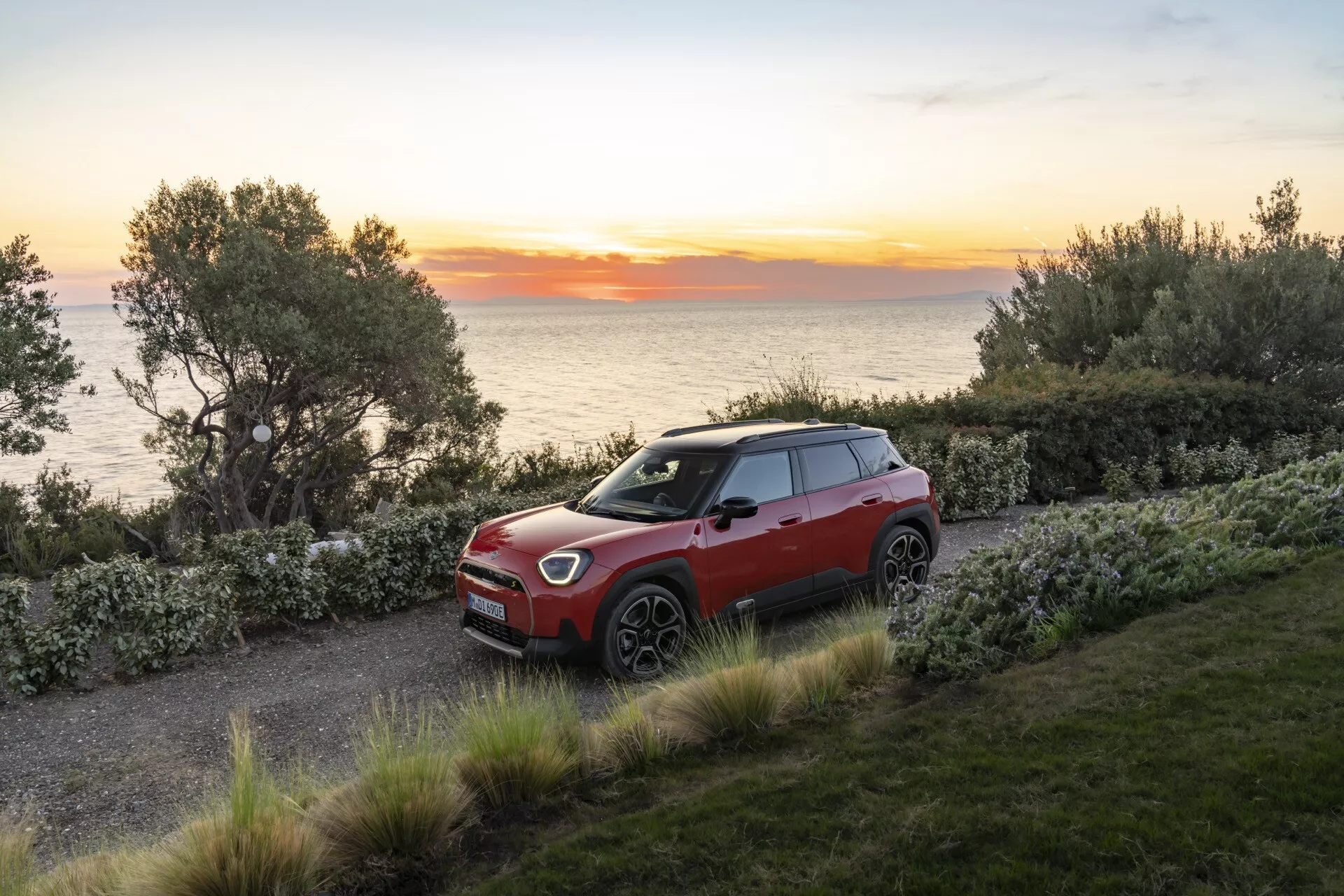 Vue à trois quarts de la Mini Aceman 2024 (J05) sur chemin de gravier présentant calandre avant, phares LED, jantes alliage multibranches, fonctionnalités avancées d'assistance à la conduite, schéma bicolore, design SUV compact, et toit ouvrant dans un cadre côtier pittoresque au lever ou coucher du soleil avec arbres, arbustes et vue sur la mer.