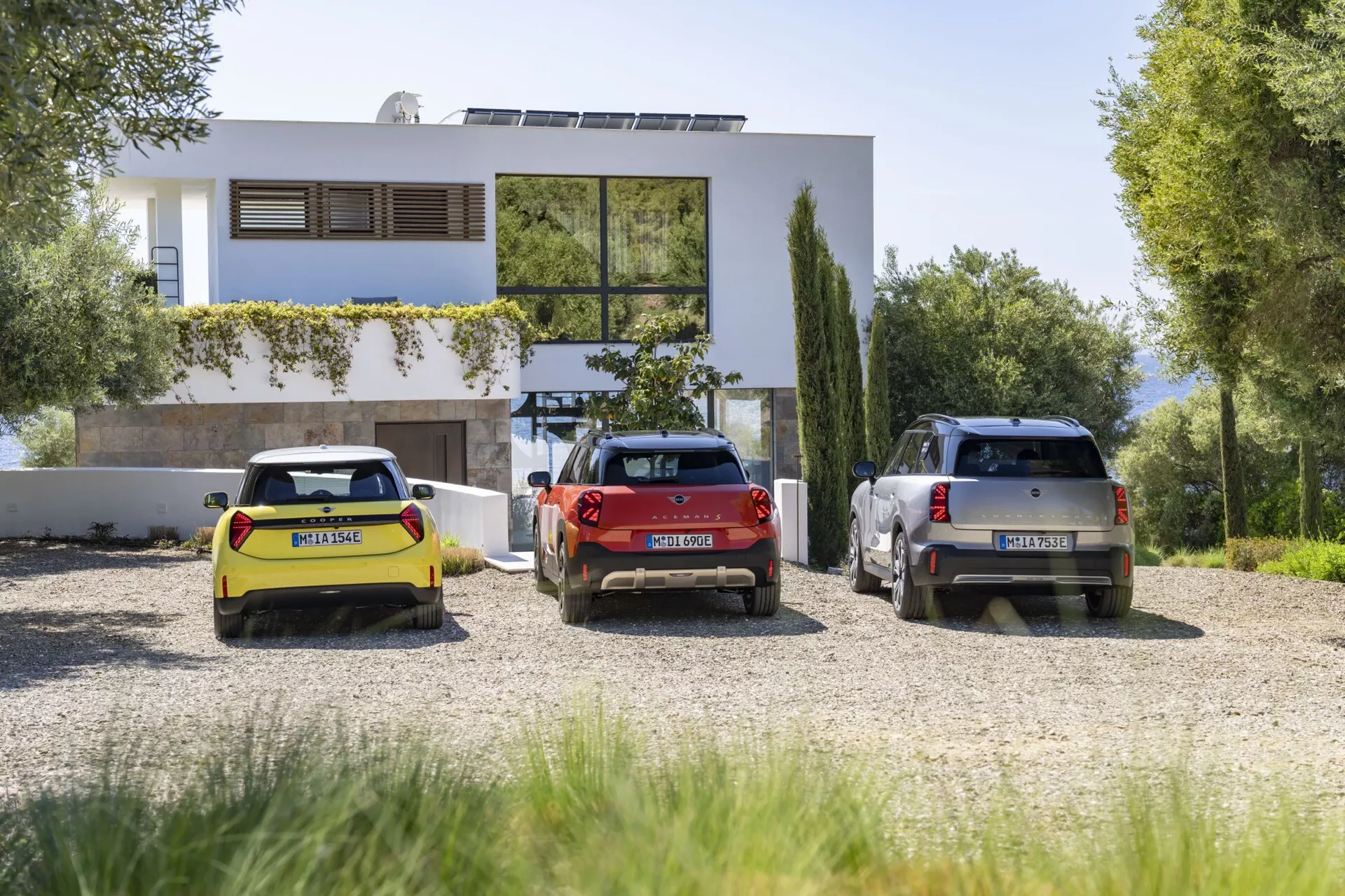 Trois modèles Mini Aceman 2024 (J05) - un cabriolet jaune, une hatchback rouge et un SUV gris - garés devant une maison moderne à deux étages parmi une verdure luxuriante, présentant des vues arrière à trois quarts et garés en parallèle sur un terrain gravillonné.