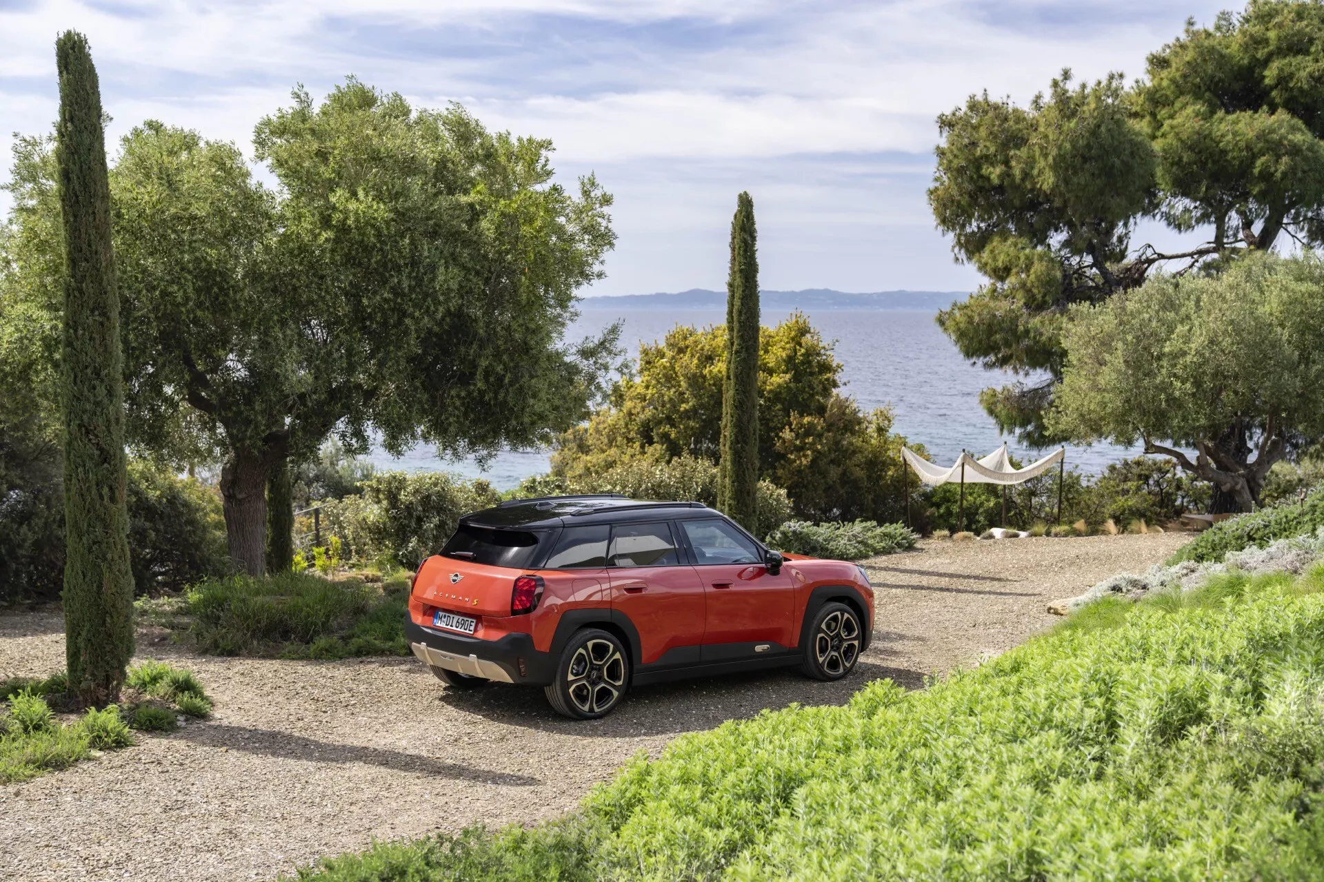 SUV compact Mini Aceman 2024 en rouge, vue arrière trois quarts garé sur un chemin de gravier dans un cadre côtier naturel avec la mer et la verdure en arrière-plan, mettant en évidence les feux arrière, les tuyaux d'échappement, le hayon avec essuie-glace, la couleur contrastante du toit et les roues en alliage.