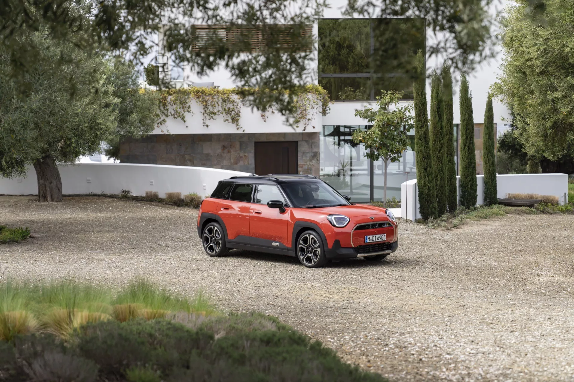 SUV compact Mini Aceman 2024 en orange vibrant avec toit noir, garé sur gravier montrant une vue de trois quarts avant contre un arrière-plan résidentiel