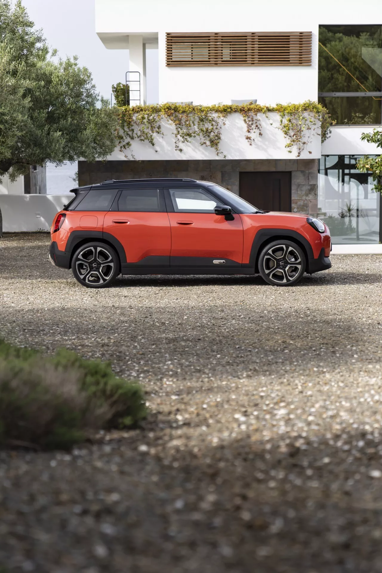 Vue de profil latéral du SUV compact Mini Aceman (J05) 2024 sur gravier, présentant une garde au sol élevée, des passages de roue proéminents, des barres de toit, des vitres à encadrement noir moderne, une carrosserie bicolore orange et noire, un moulage protecteur sur les portes et des jantes alliage